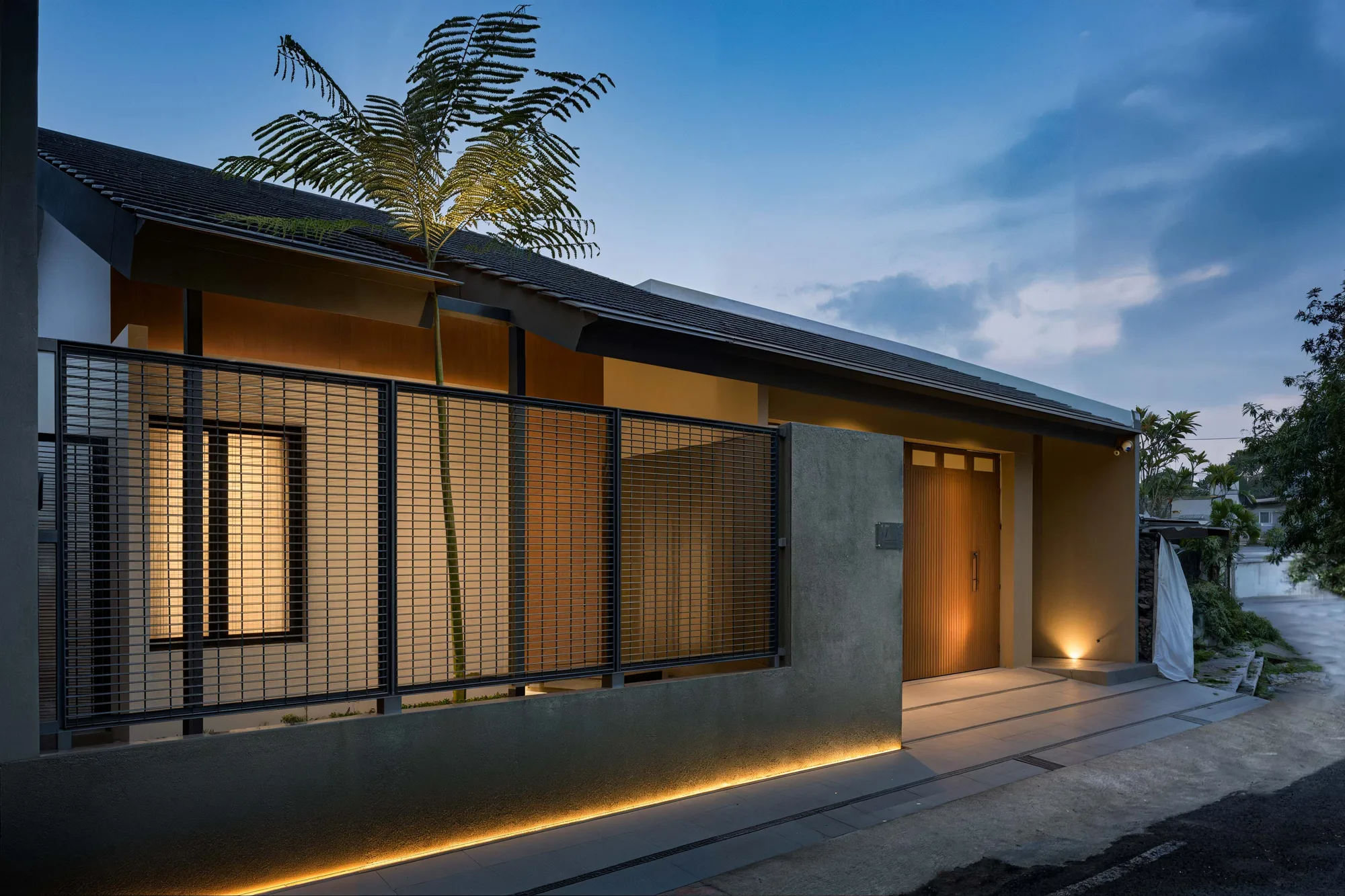 Night view of the entry facade, the timber-slatted door lit warm against grey plaster and a folding steel-mesh gate.