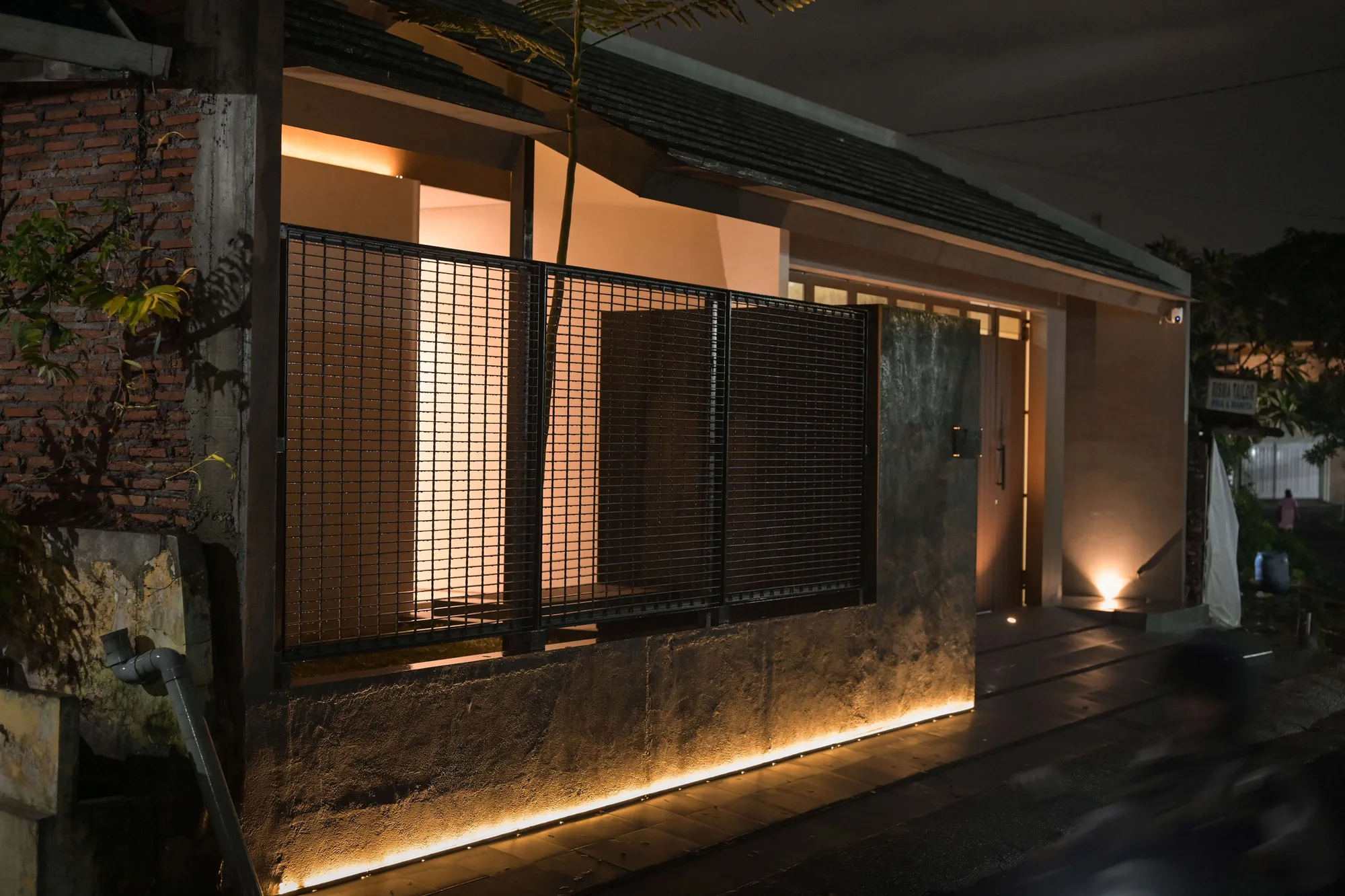 Night approach along the front pathway, warm light spilling from the mesh-screened window past the concrete gate wall to the entrance.