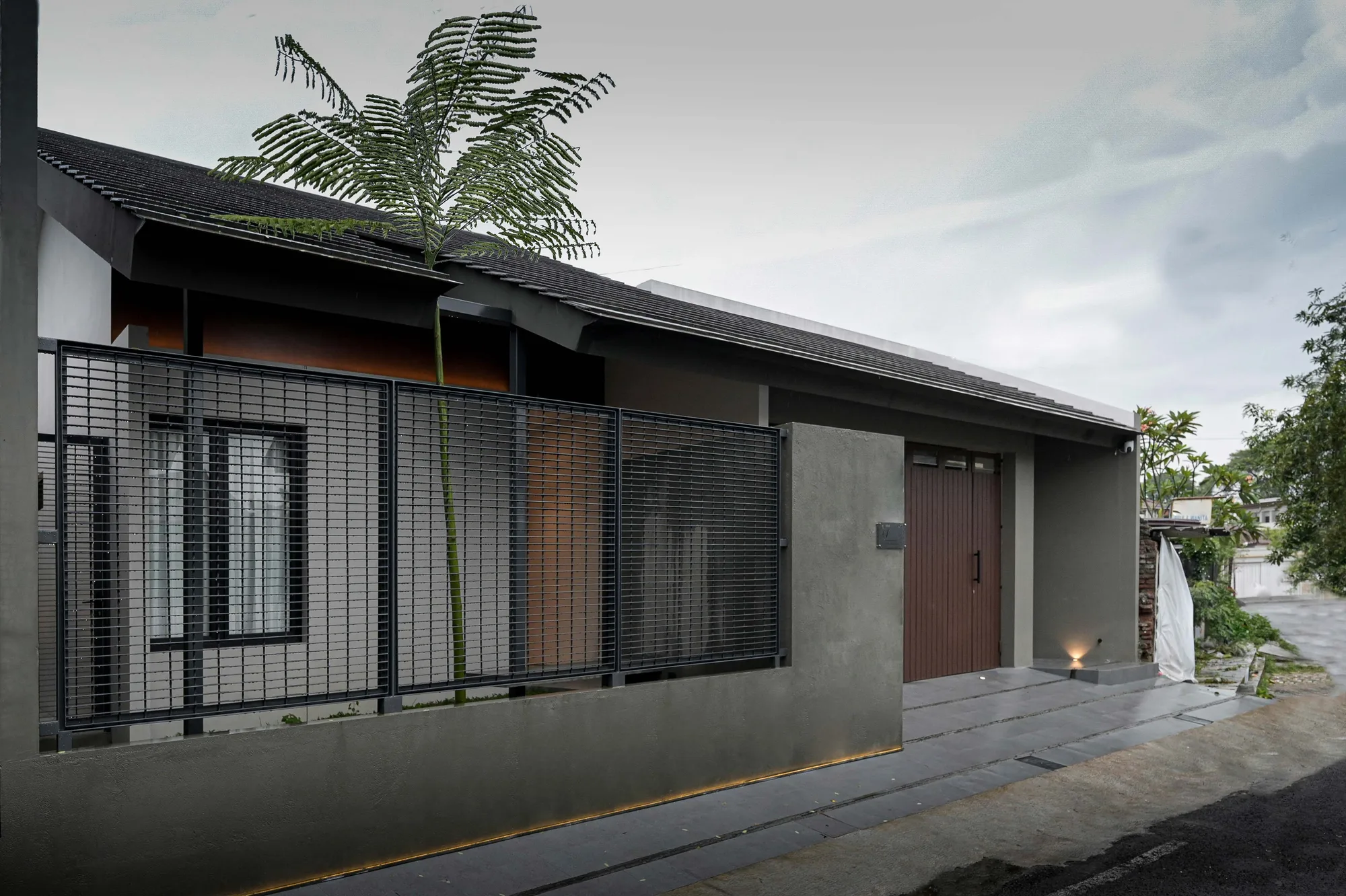 Street elevation of AL house at dusk, a slatted steel screen fronting the garage beneath a dark tiled hip roof, a tall areca palm rising past the eave.