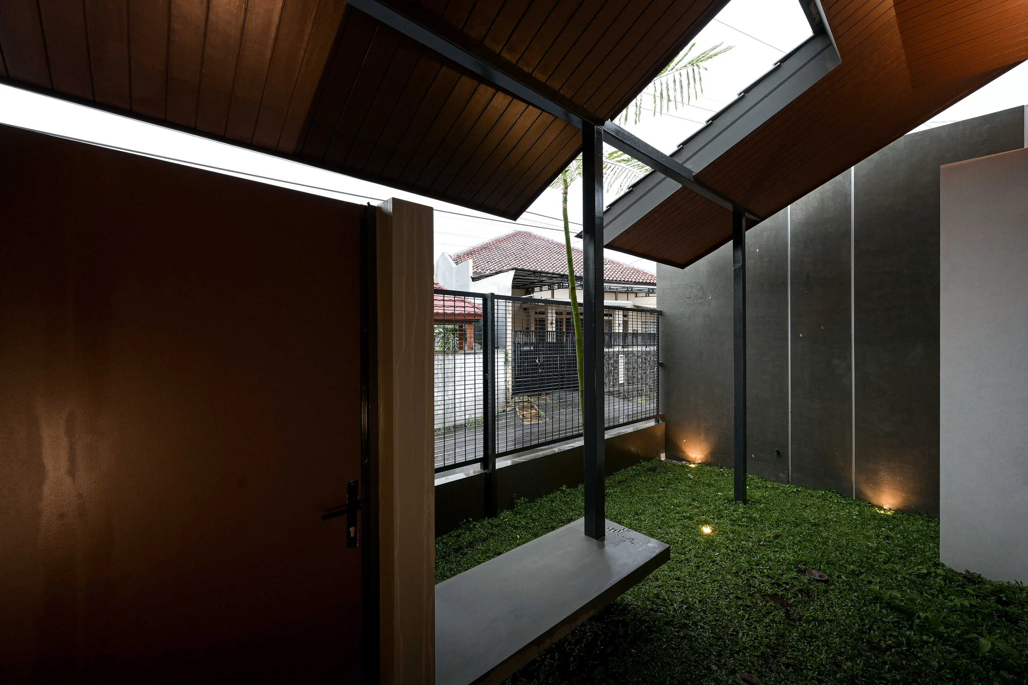 Side entry courtyard with a grass floor and angled timber-lined canopy, glimpsing the neighbouring street through a mesh fence.