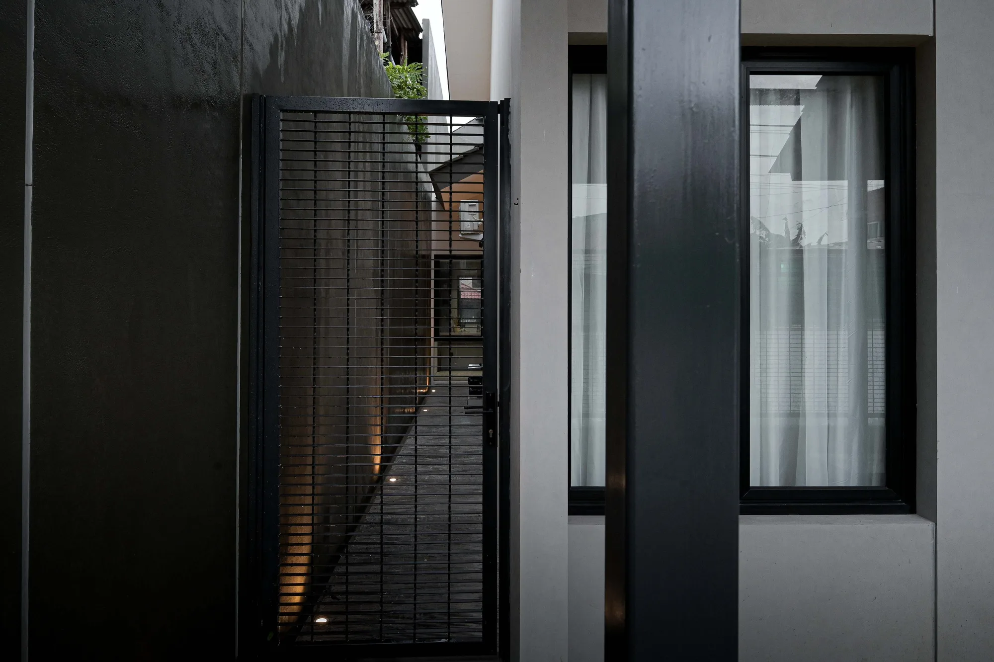 Side passage framed by a black mesh gate, glass window and dark render wall leading toward the street.