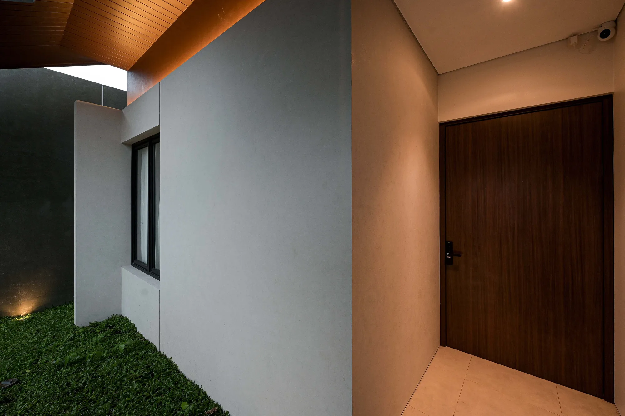 Covered walkway between the grass side yard and the timber front door, lit warm beneath an angled wood-lined soffit.