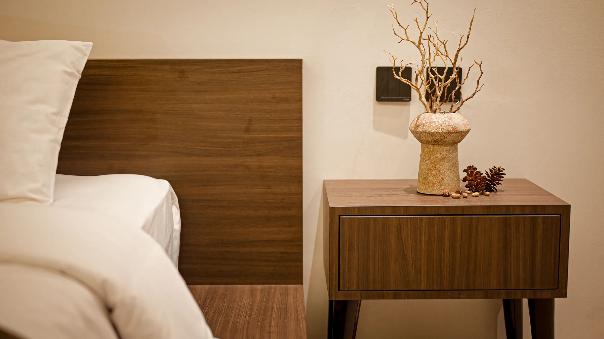Bedside table in walnut veneer holding a textured stone vase with dried branches, beside the bed's plain wood headboard.