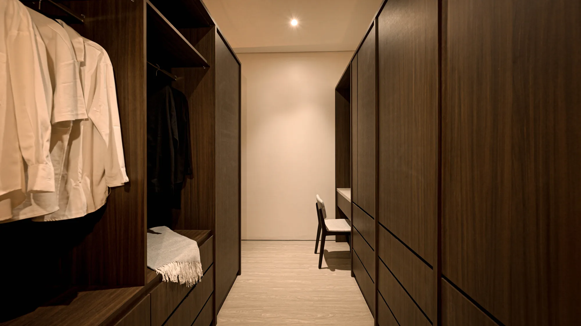 Walk-in closet corridor lined with open wardrobes, shirts hanging beside a dressing chair set at the far end.