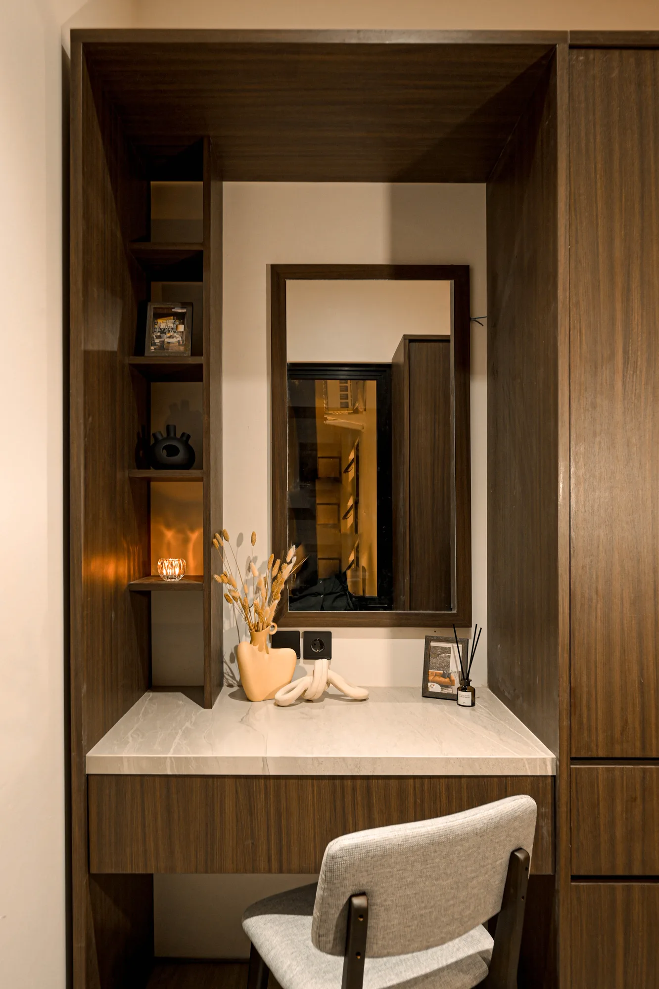 Built-in dressing nook with a mirror set into dark wood panelling, a stone vase of dried grasses on the marble counter.
