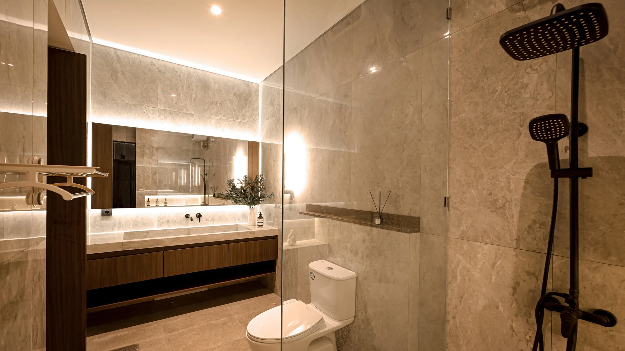 Primary bathroom with a floating walnut vanity, backlit mirror, and glass-walled shower in beige stone tile.