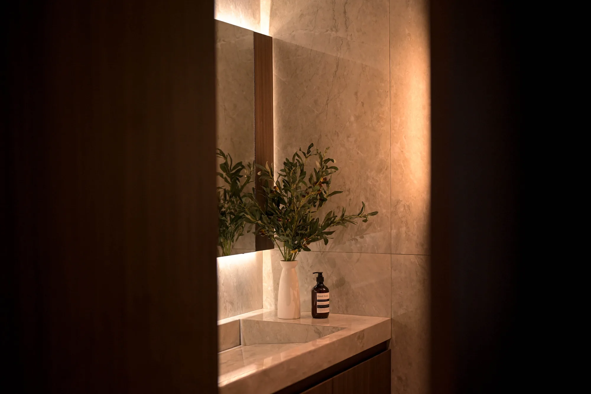 Bathroom vanity seen through a cracked door, an olive branch in a ceramic vase beside the hand soap on the stone counter.