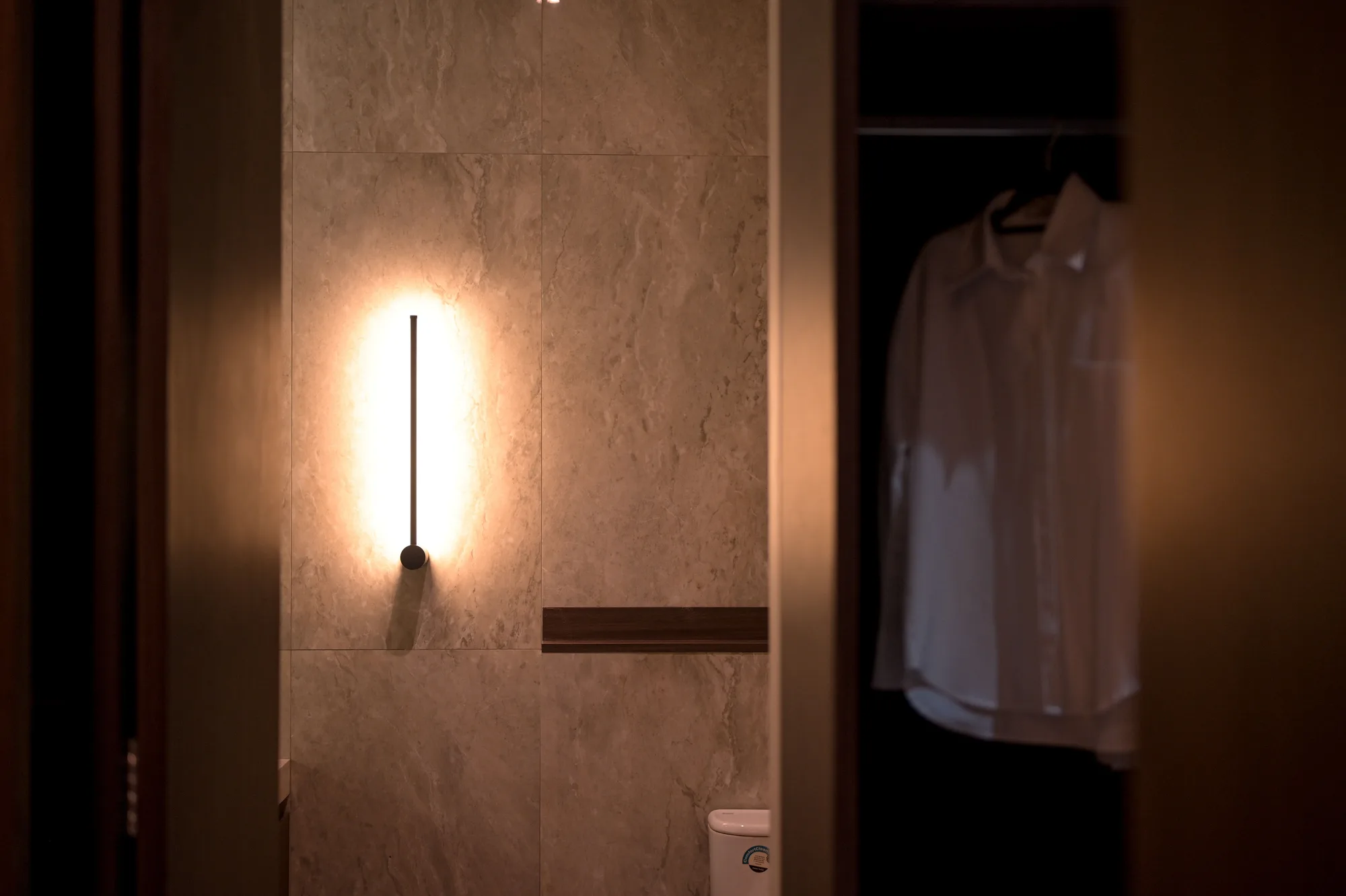 Vertical wall sconce glowing against stone tile between the toilet and an adjoining wardrobe of hanging shirts.