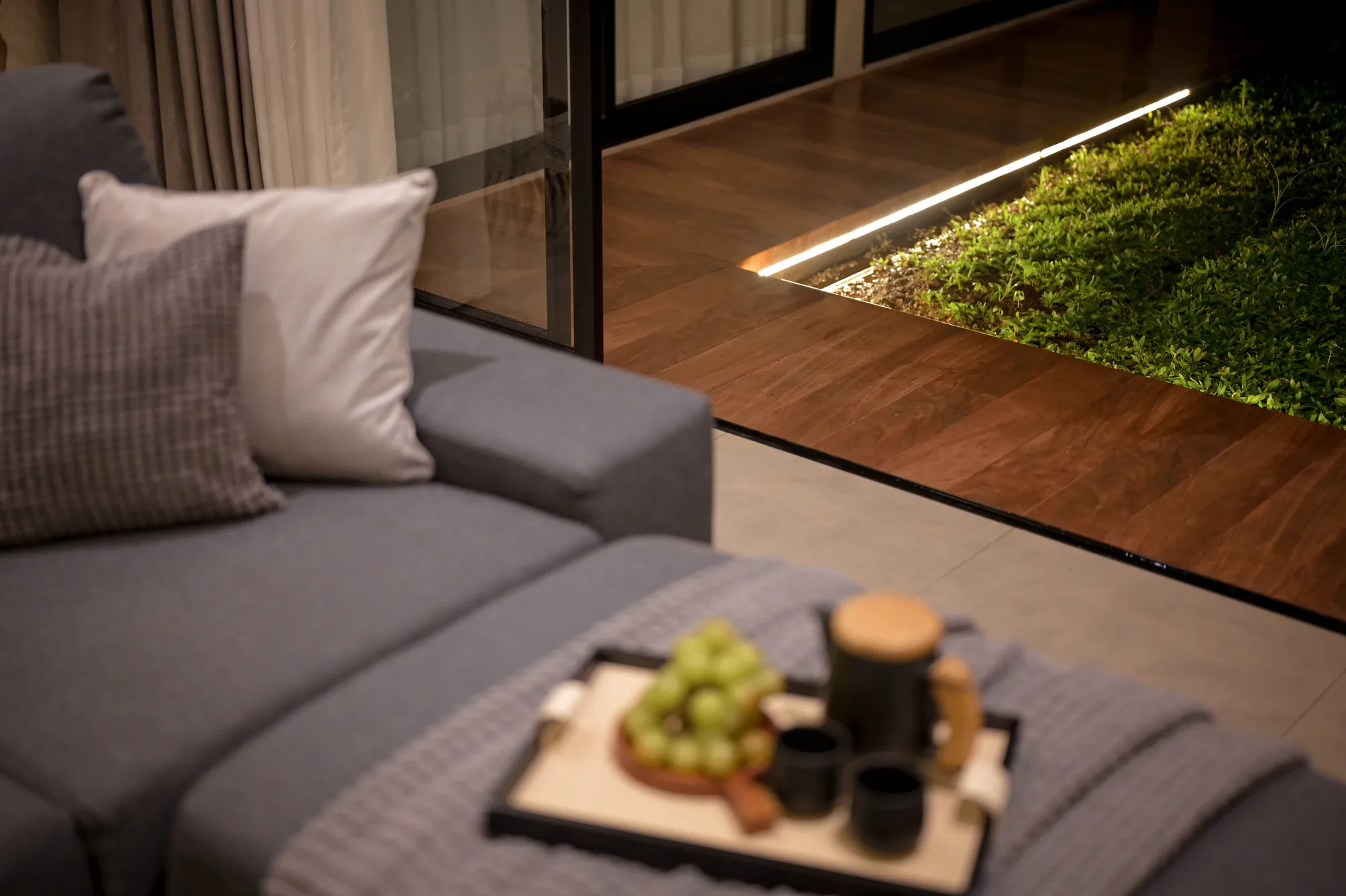 Outdoor lounge with a grey sofa and cushions beside sliding glass doors, a strip light tracing the edge of a planted garden bed at night.