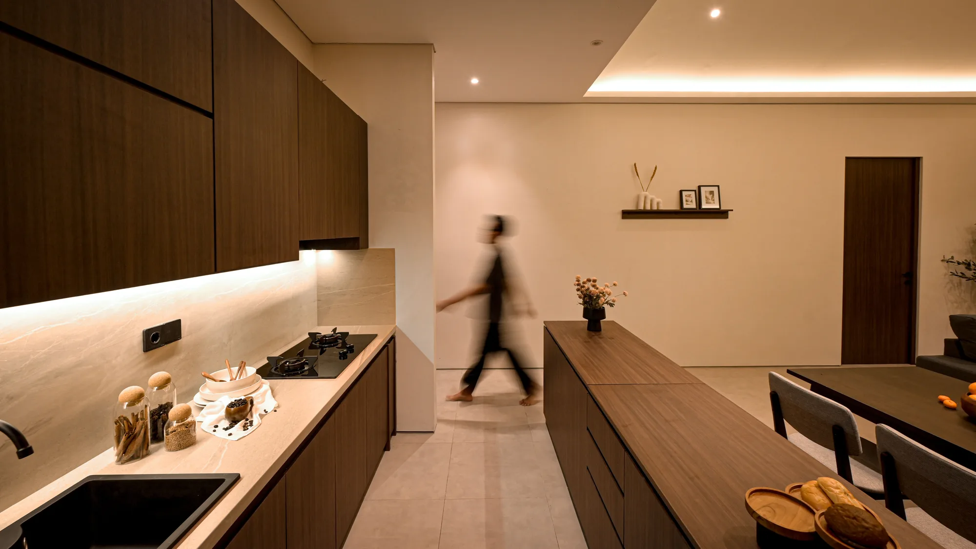 Kitchen counter in walnut cabinetry, a figure passing blurred in motion toward the dining area beyond.