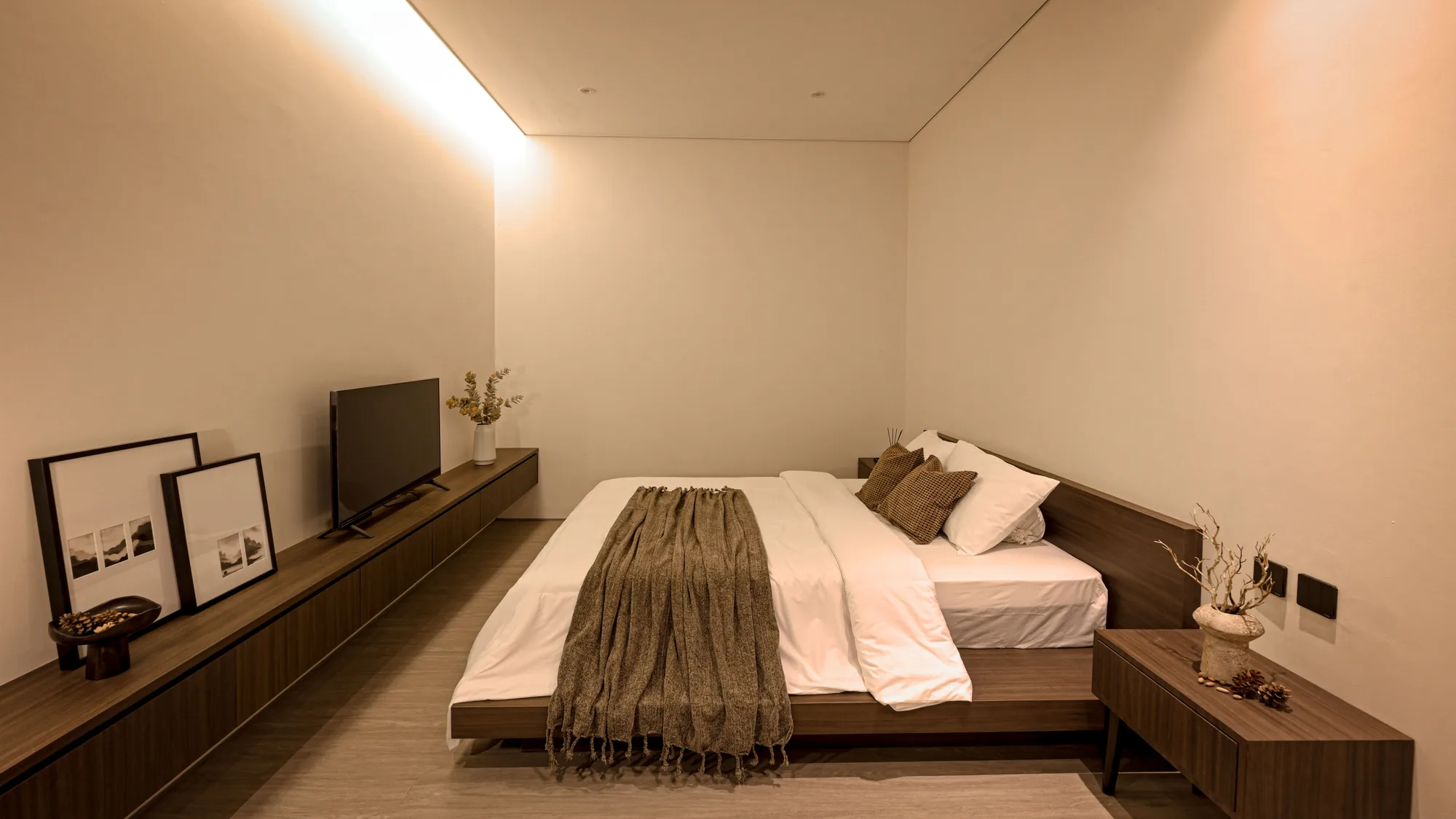 Bedroom with a low platform bed dressed in a woven throw, facing a floating console holding a television and framed photographs.