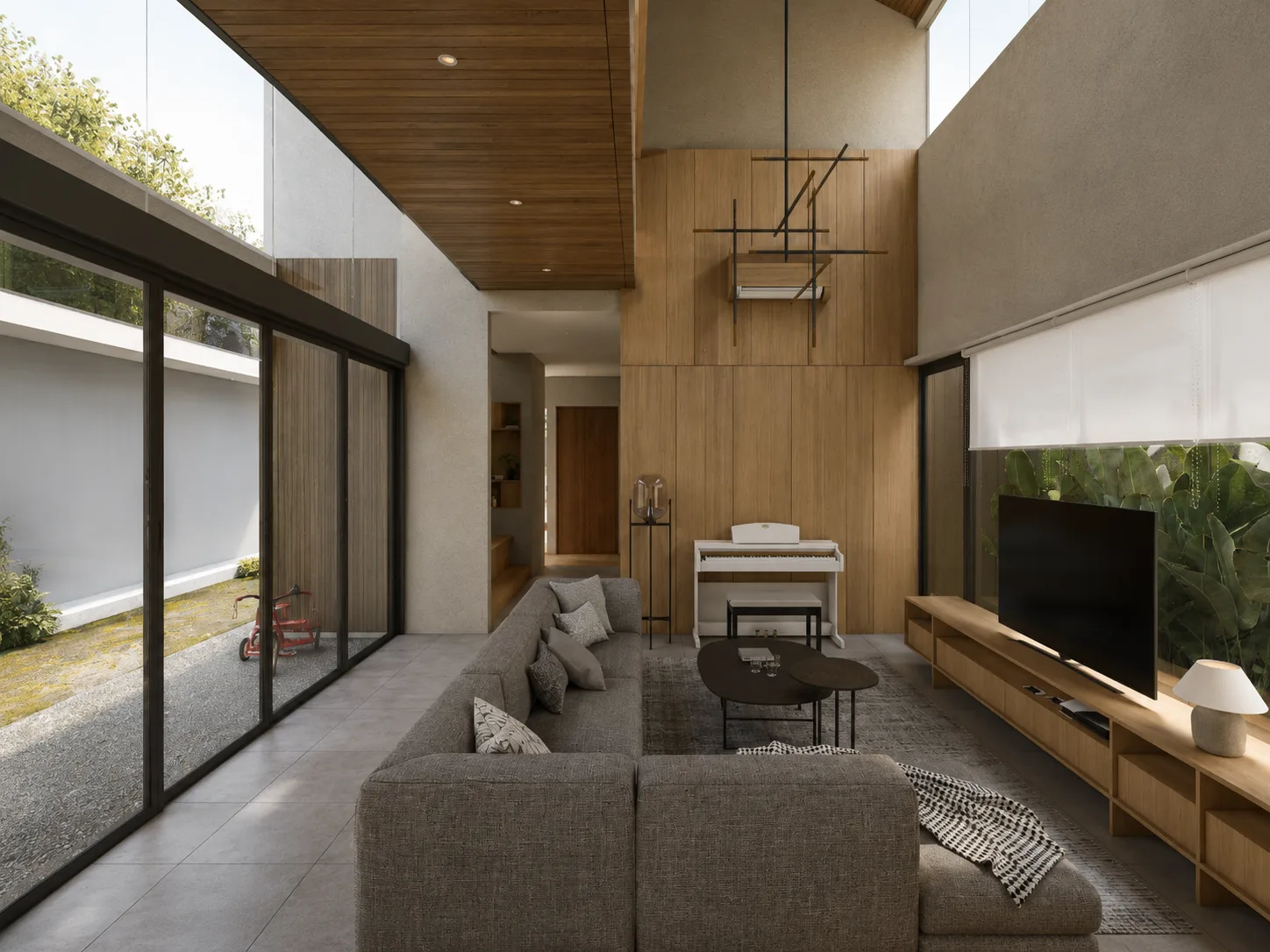 Living room corner with a grey sectional sofa and timber TV console, glazed walls looking onto a gravel courtyard with a tricycle.