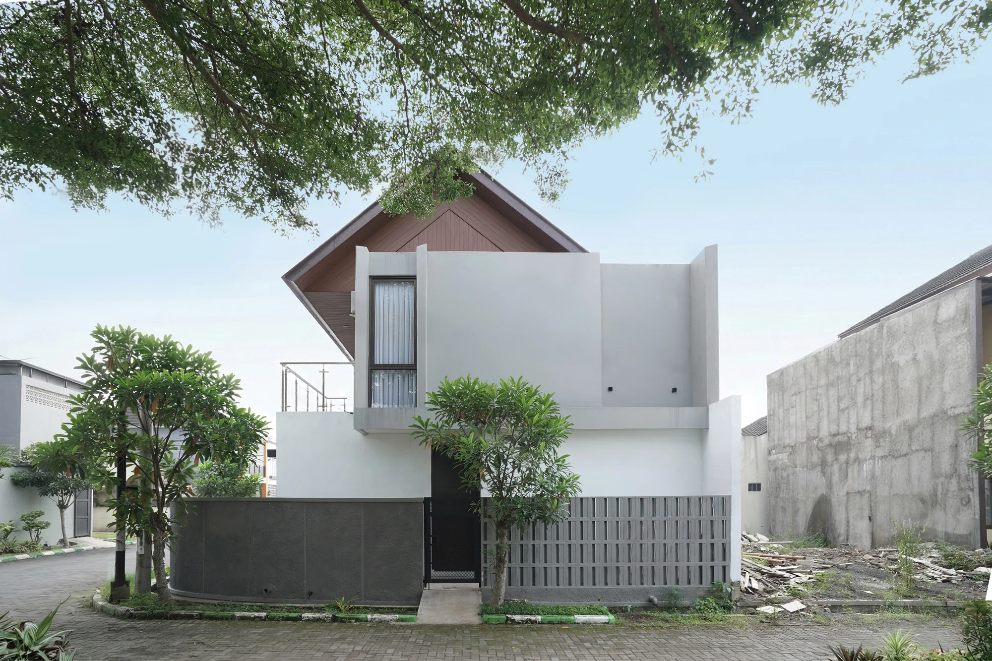 A close view of Hook House's gable end, its warm wood-lined underside floating above plain white walls, framed by an overhanging street tree and the dark perforated fence below.