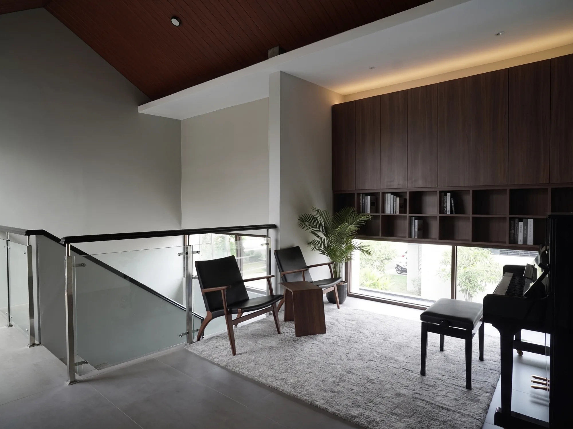 An upstairs landing opens into a piano room, its sloped wood ceiling and walnut bookshelf wall overlooking dark leather lounge chairs and a glass balustrade above the stair.