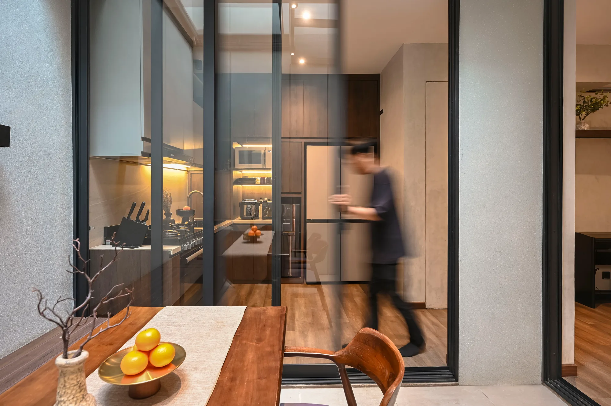 A figure moving through the kitchen, seen past the dining table and its bowl of citrus fruit.