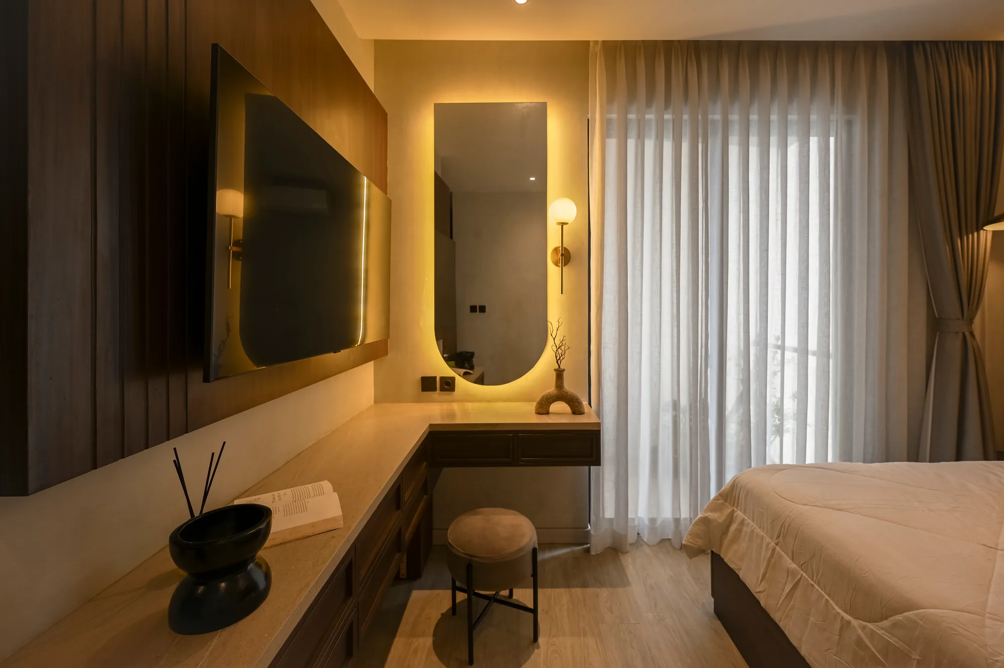 Bedroom television and backlit mirror set into a wood-panelled wall beside sheer curtains.