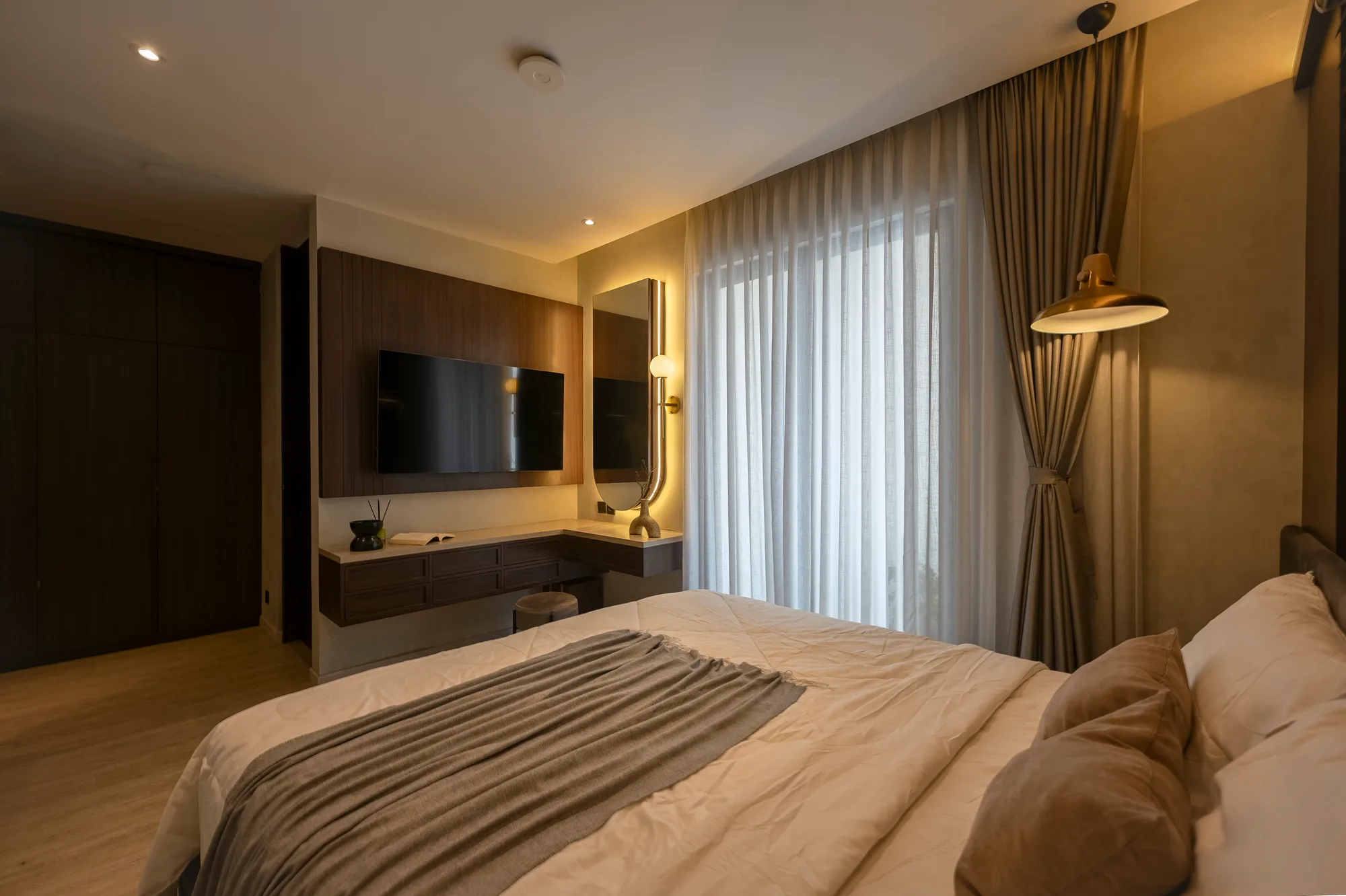 Bedroom viewed from the foot of the bed, wardrobe, television console and brass pendant lamp along one wall.