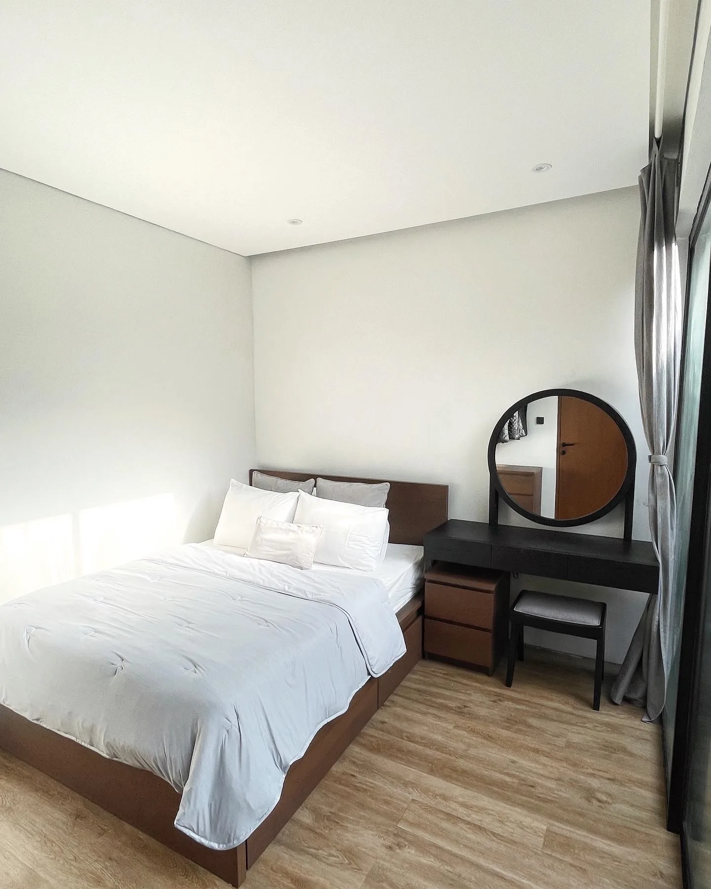 A bedroom with white linens on the bed, a round mirror standing on a dark dressing table.