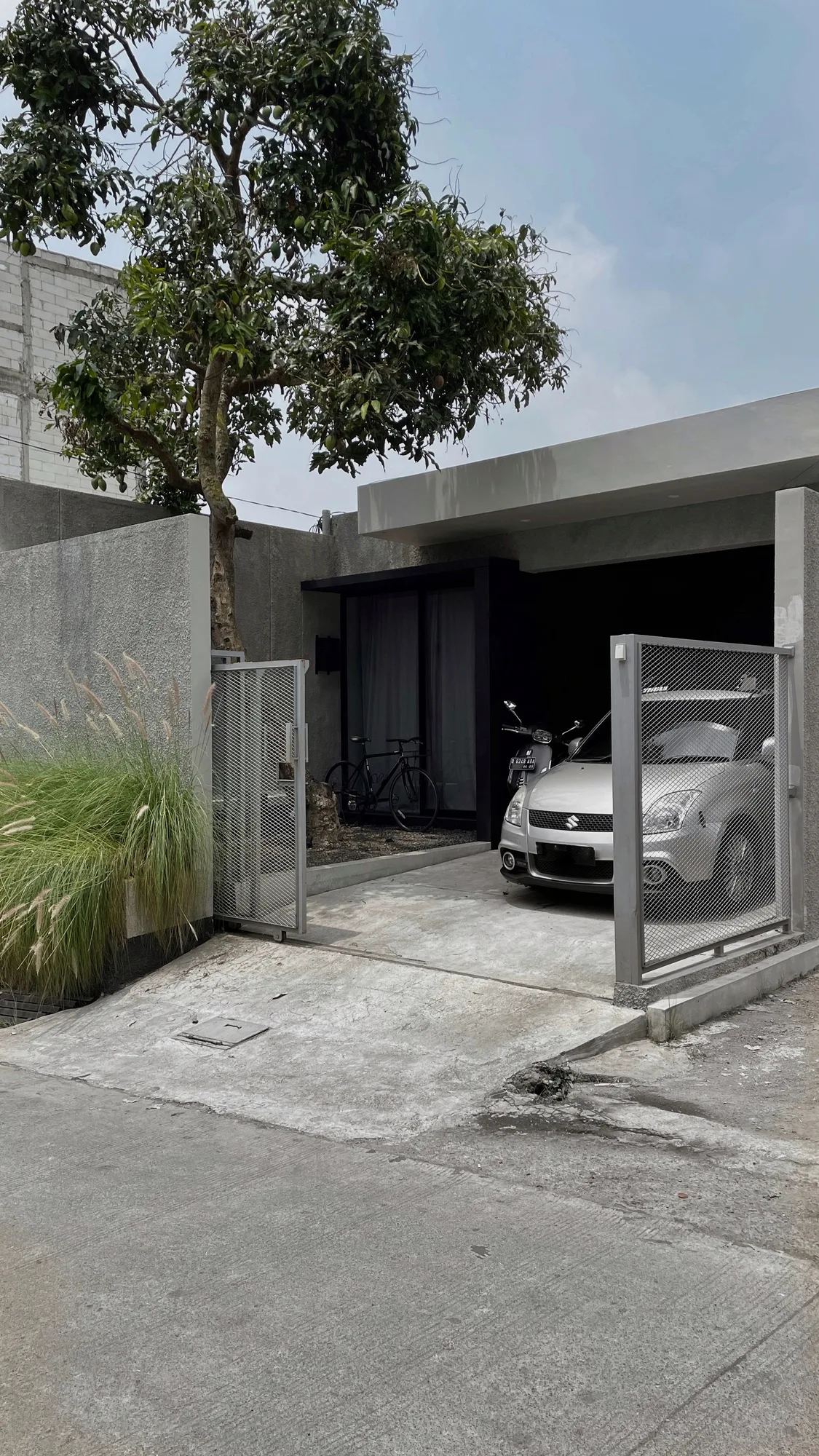 The carport by day, mesh gate open to a silver car and scooter, a mango tree overhead.
