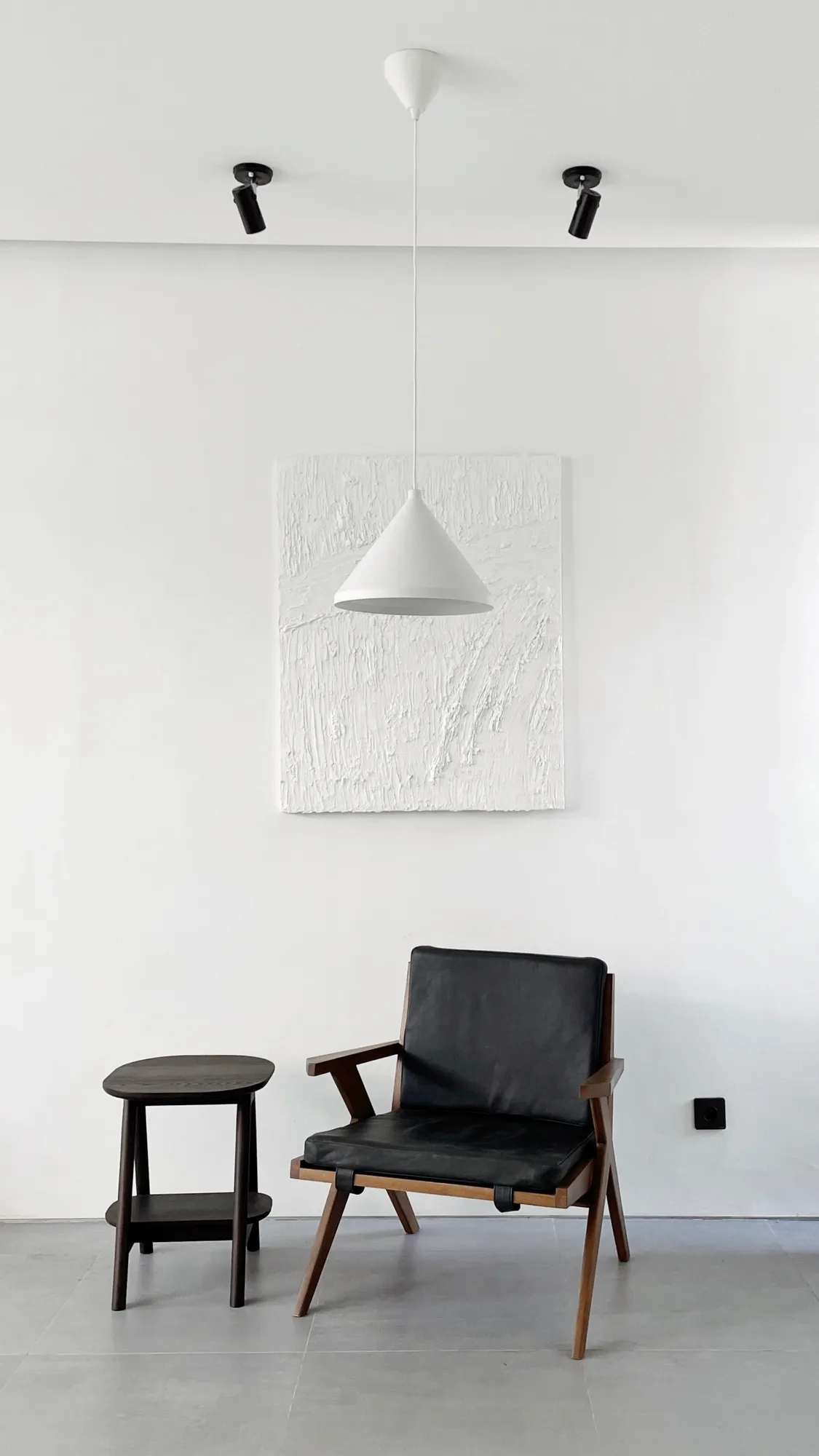 A corner with a white textured canvas, a cone pendant lamp, and a black leather lounge chair beside a side table.