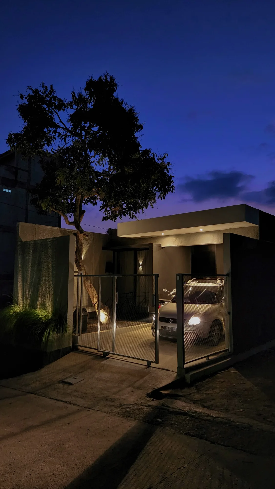 The carport at dusk, gate lit and car headlights on, a mango tree silhouetted against the evening sky.