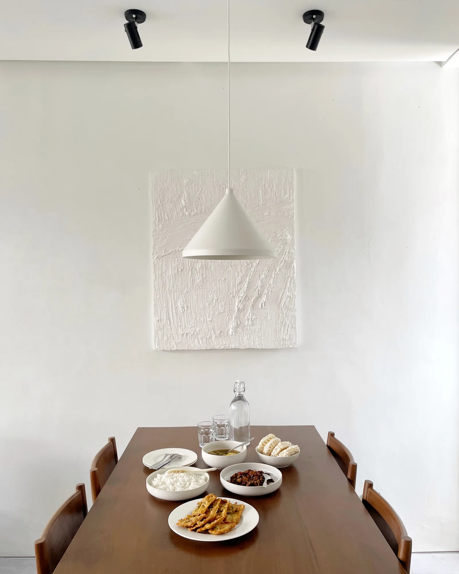 A dining table set with rice, side dishes and a bottle of water, beneath a cone pendant and a textured white canvas.