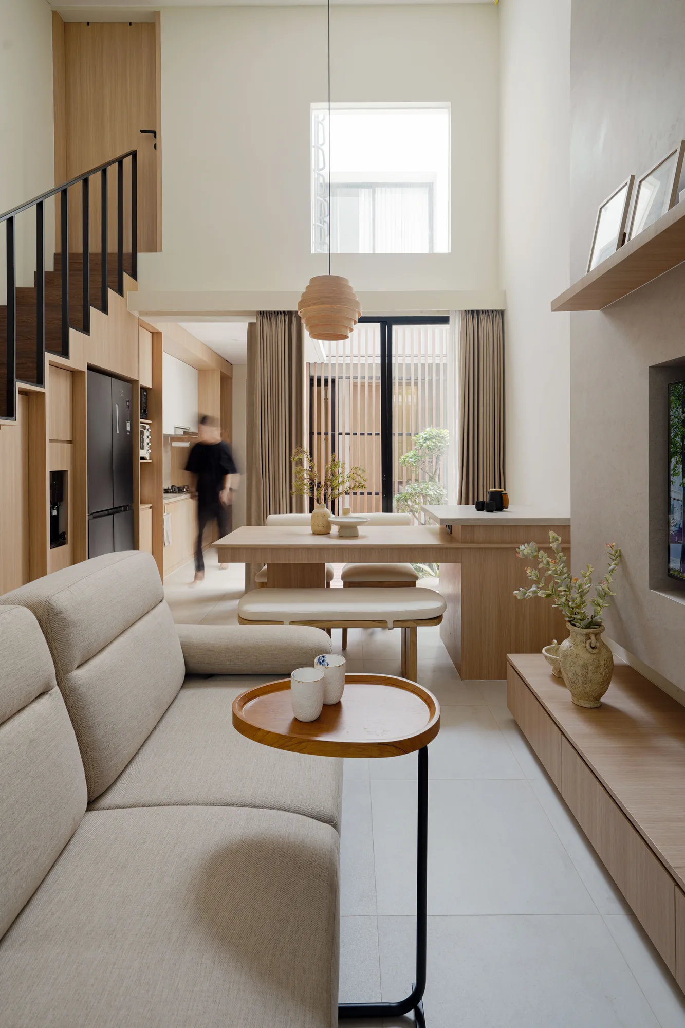 View through the double-height living room toward the courtyard, with a timber dining table and black-framed stair.
