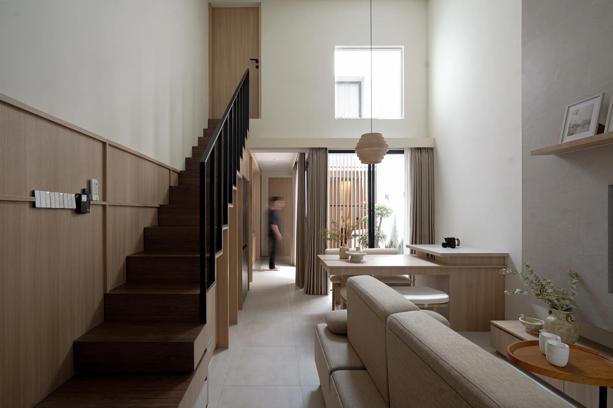Double-height living room seen from the stair, with a pale sofa, timber dining table and courtyard beyond.