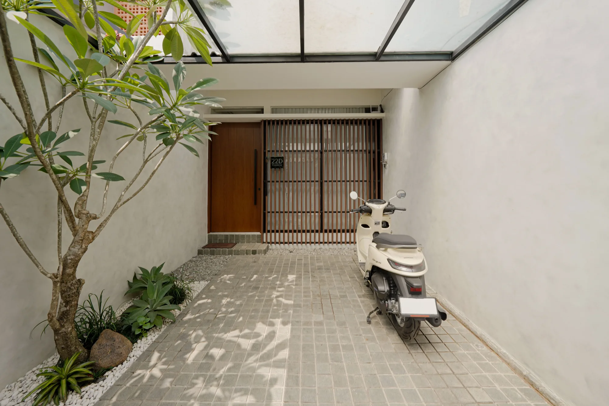 Carport garden with a frangipani tree, a timber-slat entry screen and a scooter parked on grey pavers.