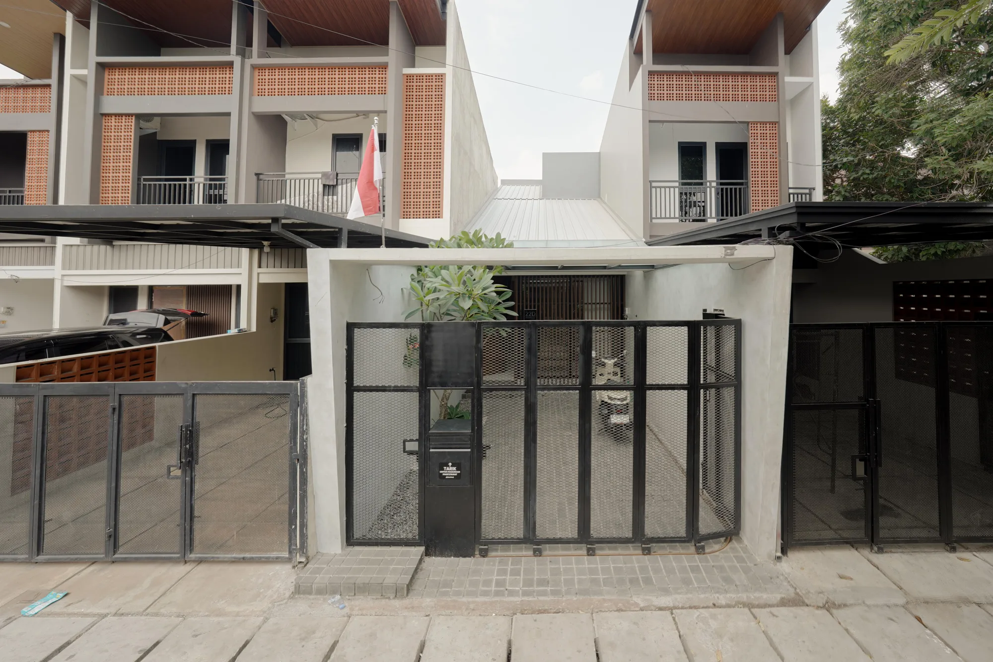Street facade of Prada House, a black perforated steel gate beneath a pale concrete carport, framed by neighboring homes.