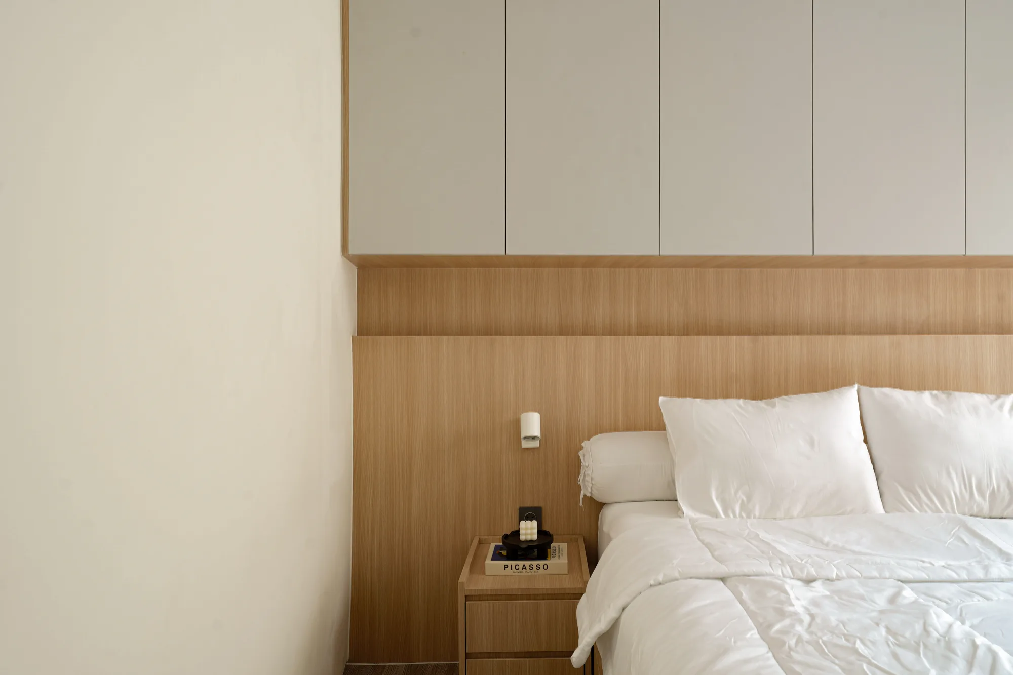 Bedroom corner with white bedding, pale timber headboard panelling and overhead cabinets.