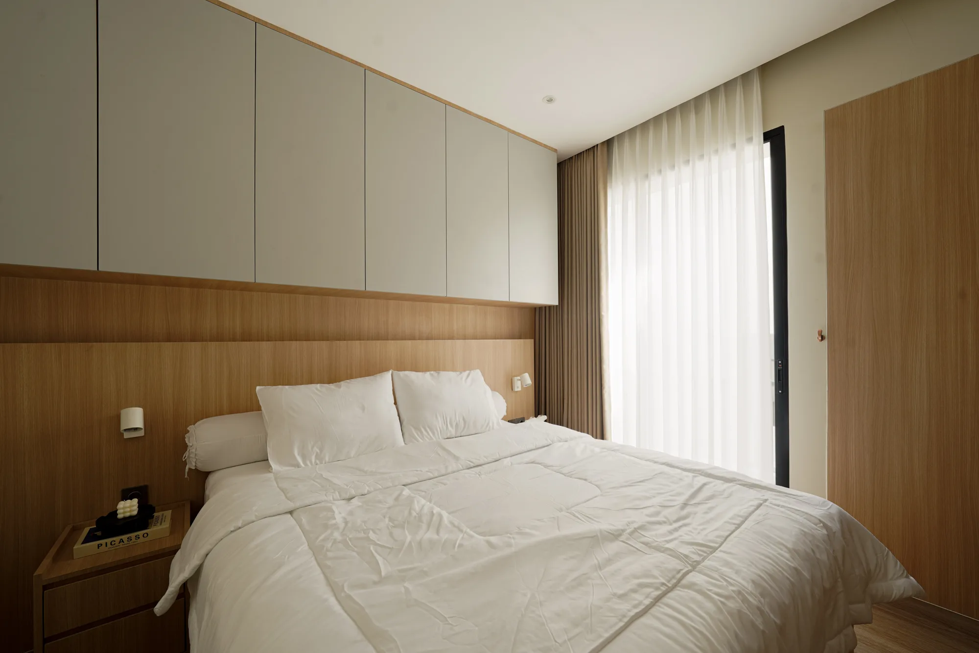 Bedroom with white bedding, full-height timber cabinetry and sheer curtains filtering daylight.
