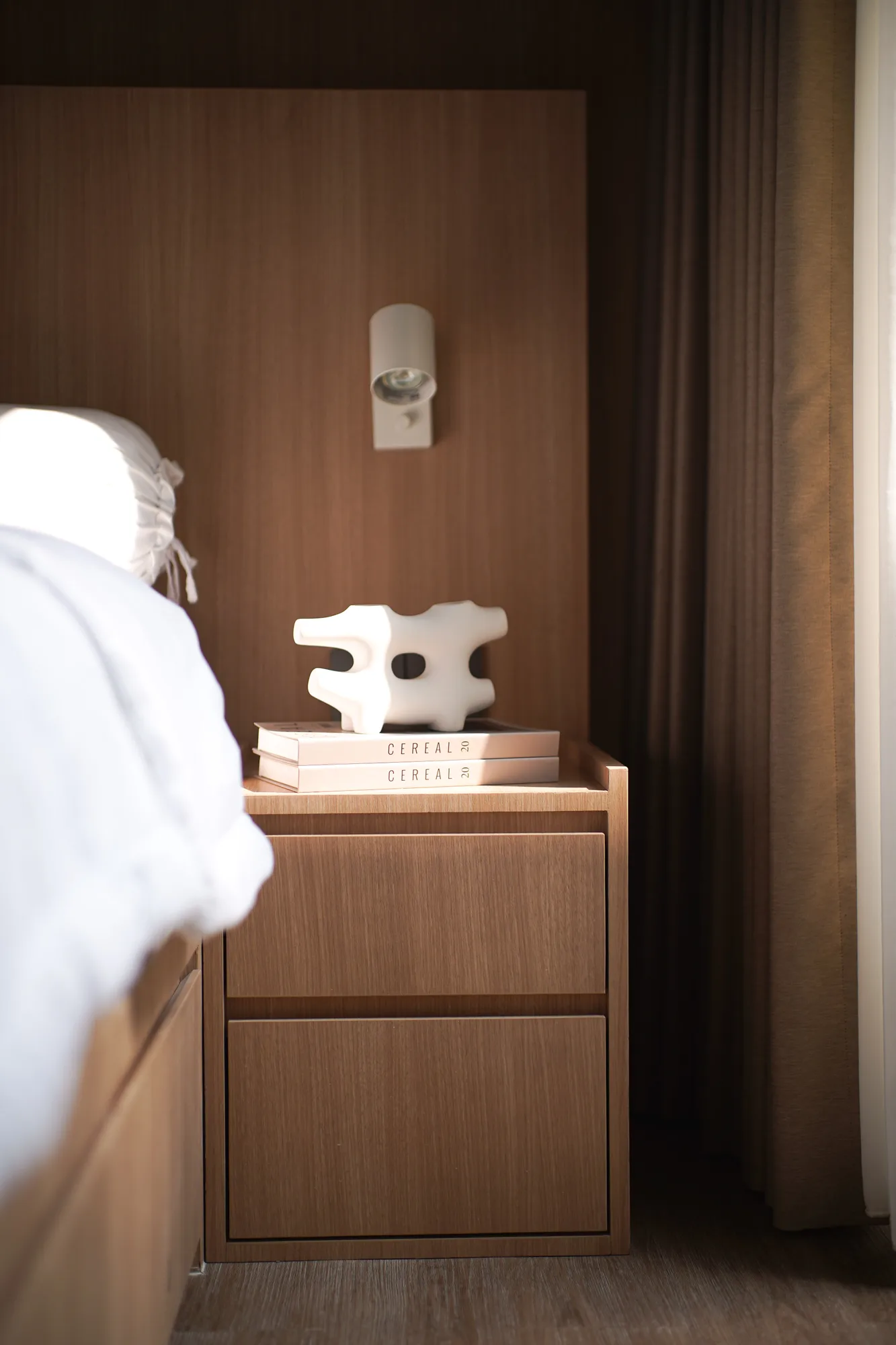 Bedside table in pale timber, topped with art books and a white ceramic sculpture beside drawn curtains.