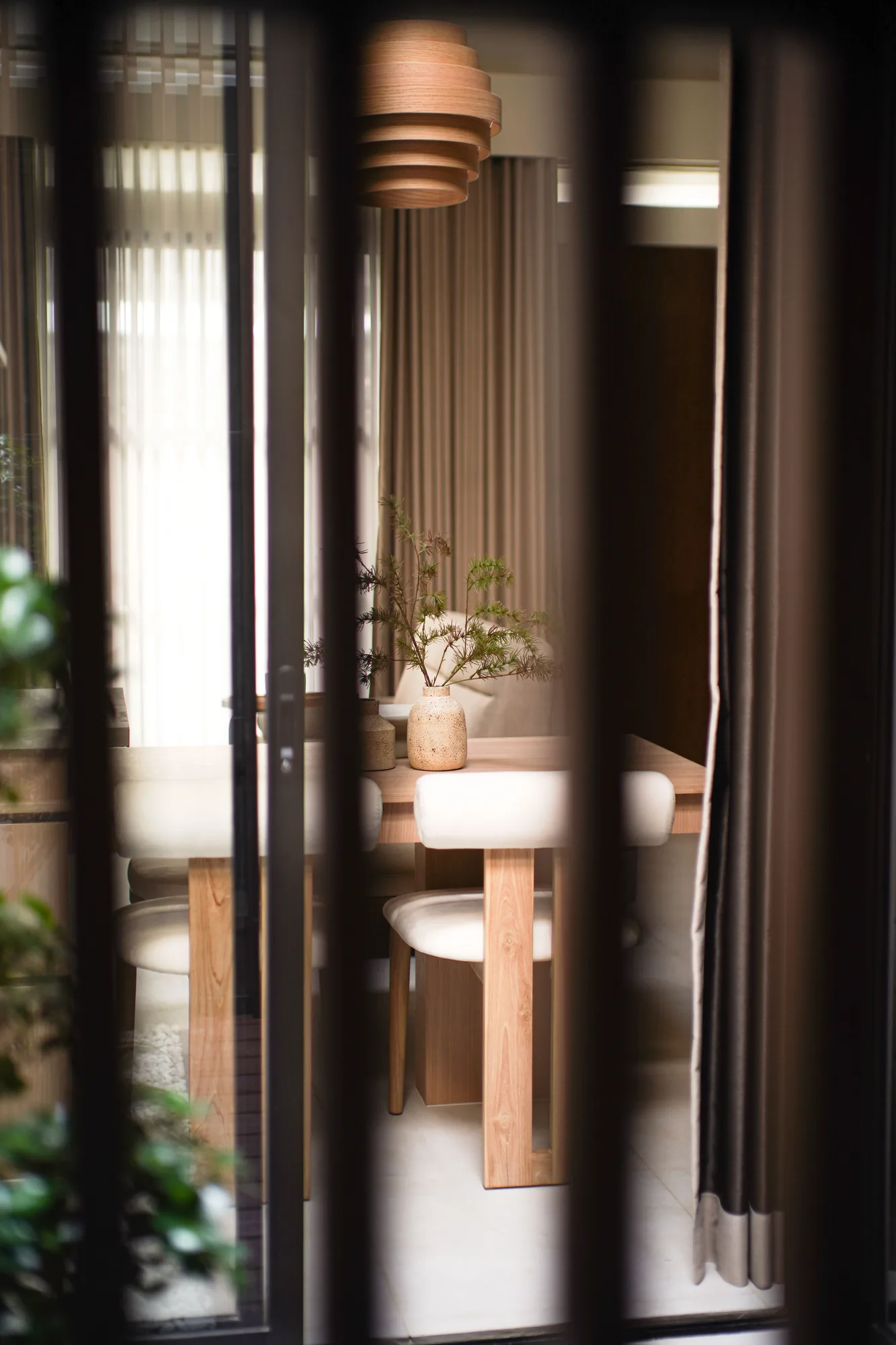 Timber pendant light and dining table seen through dark vertical courtyard frames.