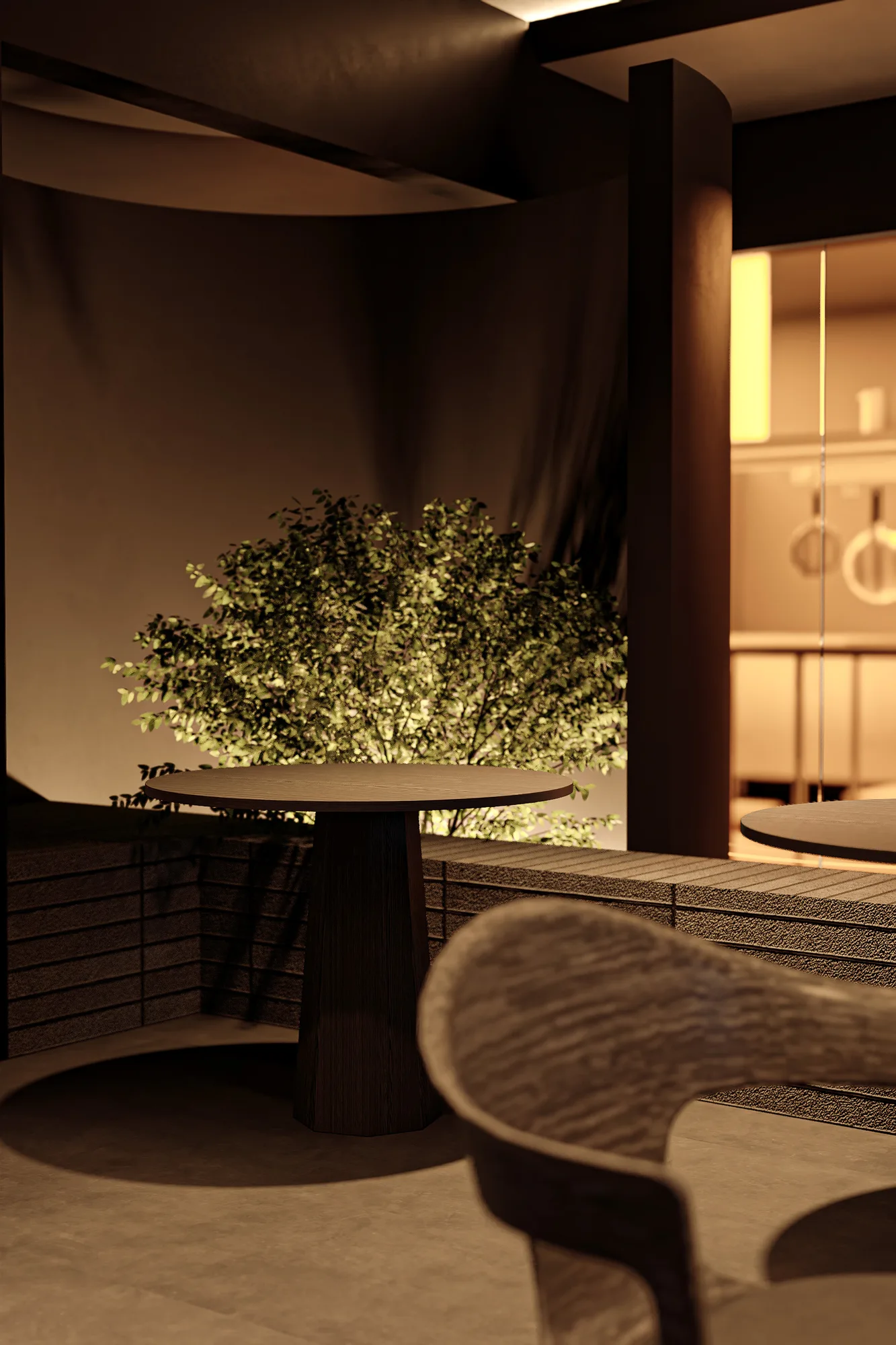 A round wood table on the terrace, the kitchen visible through the glass wall behind it.