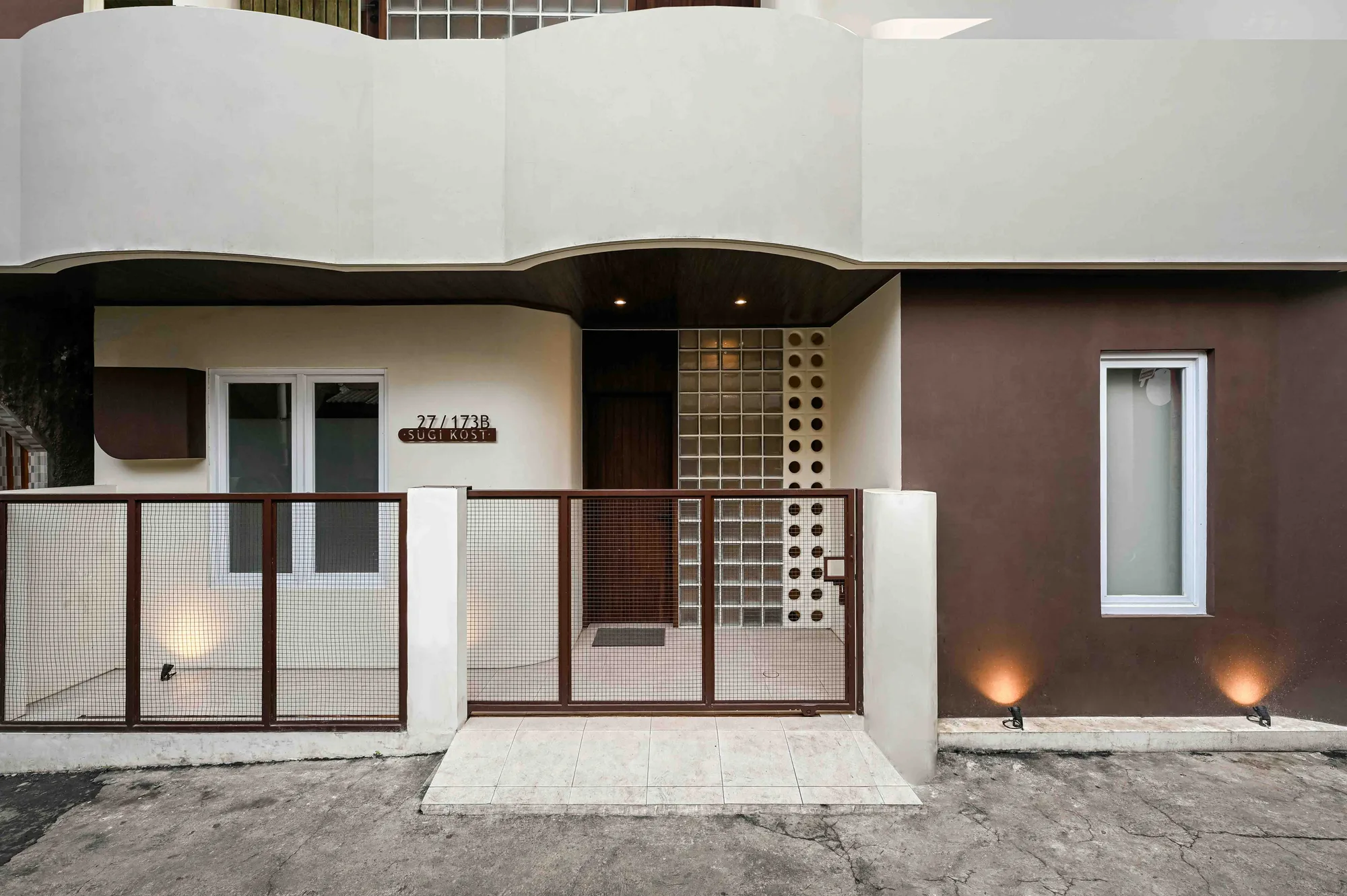 Sugi Kost's street facade with its wire-mesh gate closed and glass-block entrance wall lit beneath a curved white overhang.