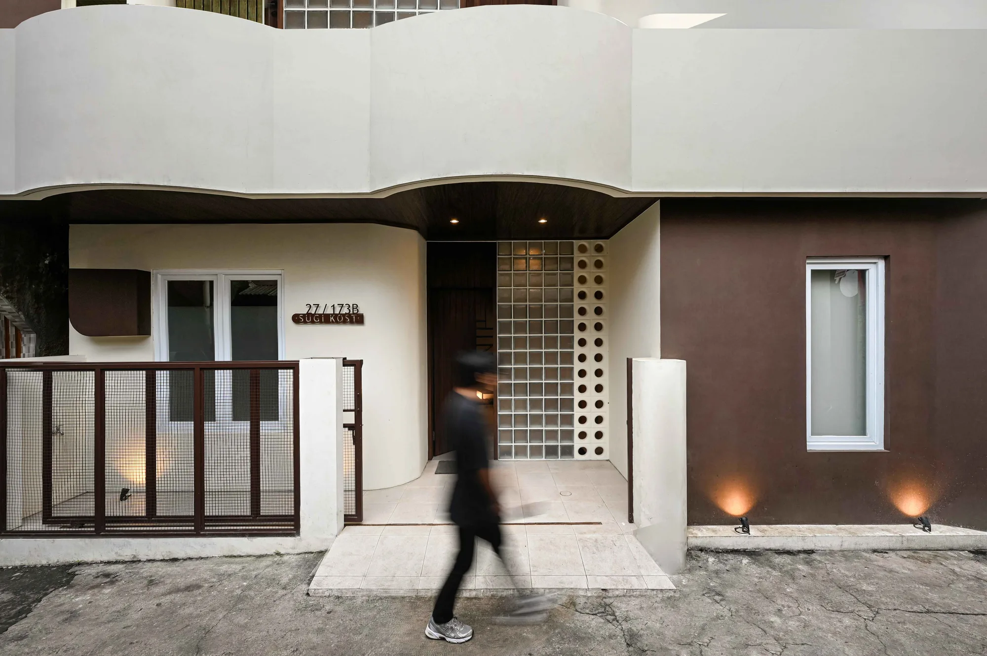 A resident crossing the gated entrance courtyard beneath the curved white overhang of Sugi Kost's street facade.