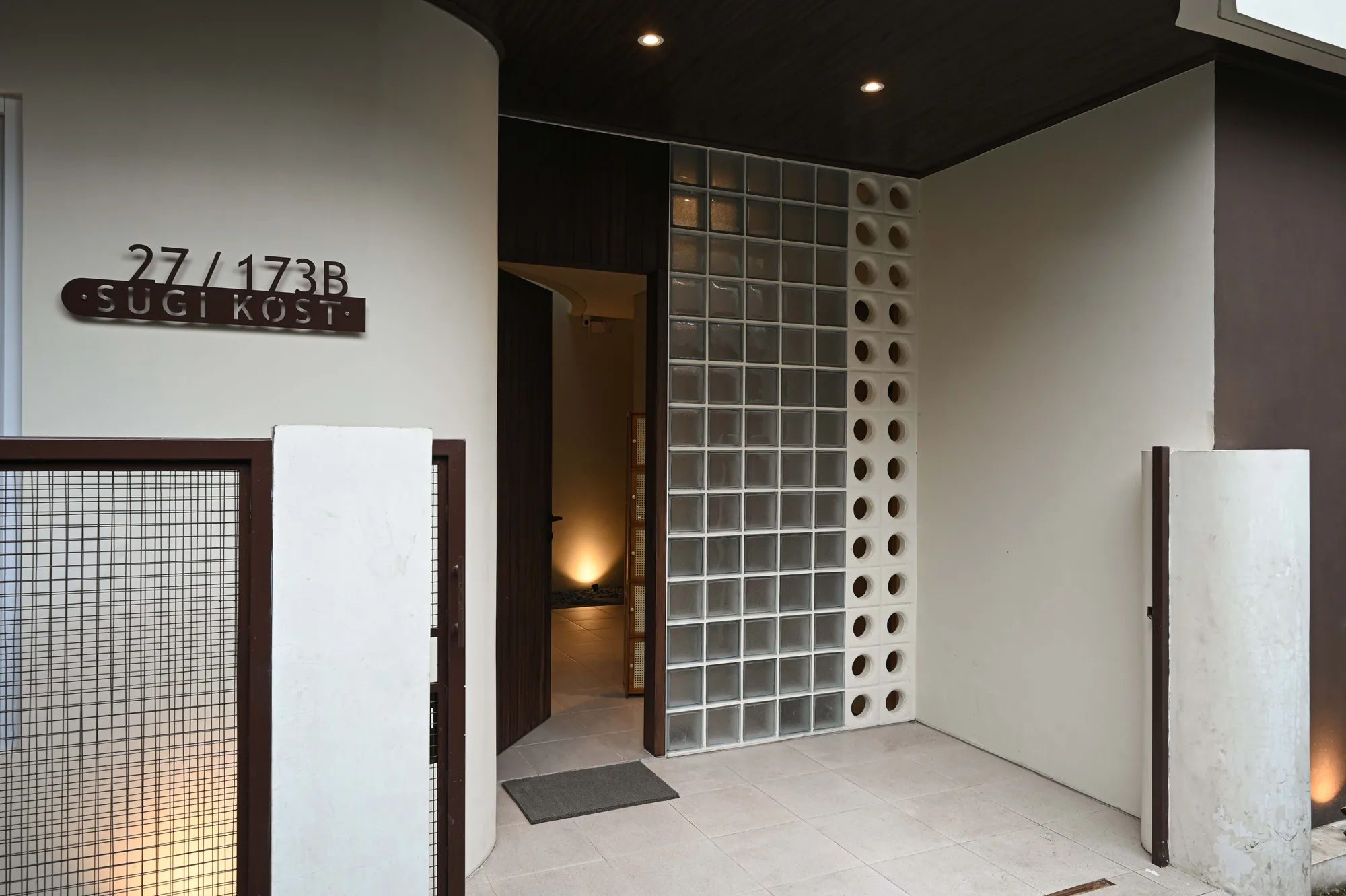 The entrance vestibule where a glass-block wall and a perforated concrete column meet beside the Sugi Kost signage and open front door.