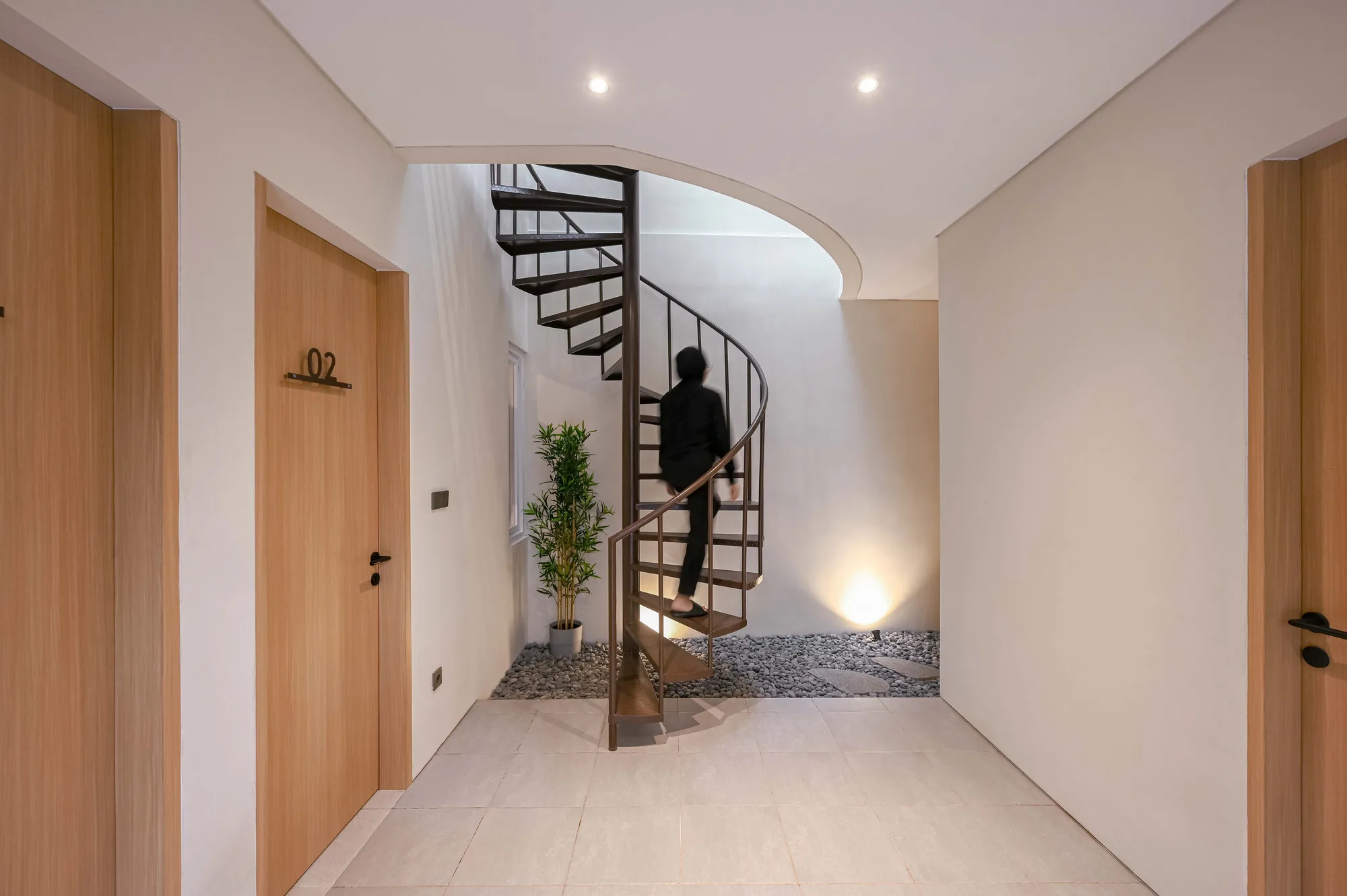 A resident descending the dark spiral stair that threads through the vertical spine, with room 02's door at the base.
