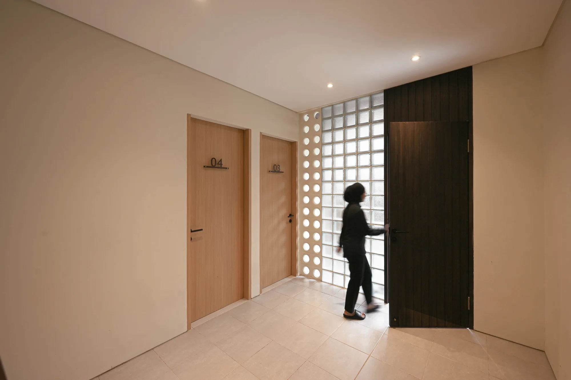 Corridor where a resident opens the dark front door beside a full-height glass-block wall, doors 03 and 04 lining the hall.