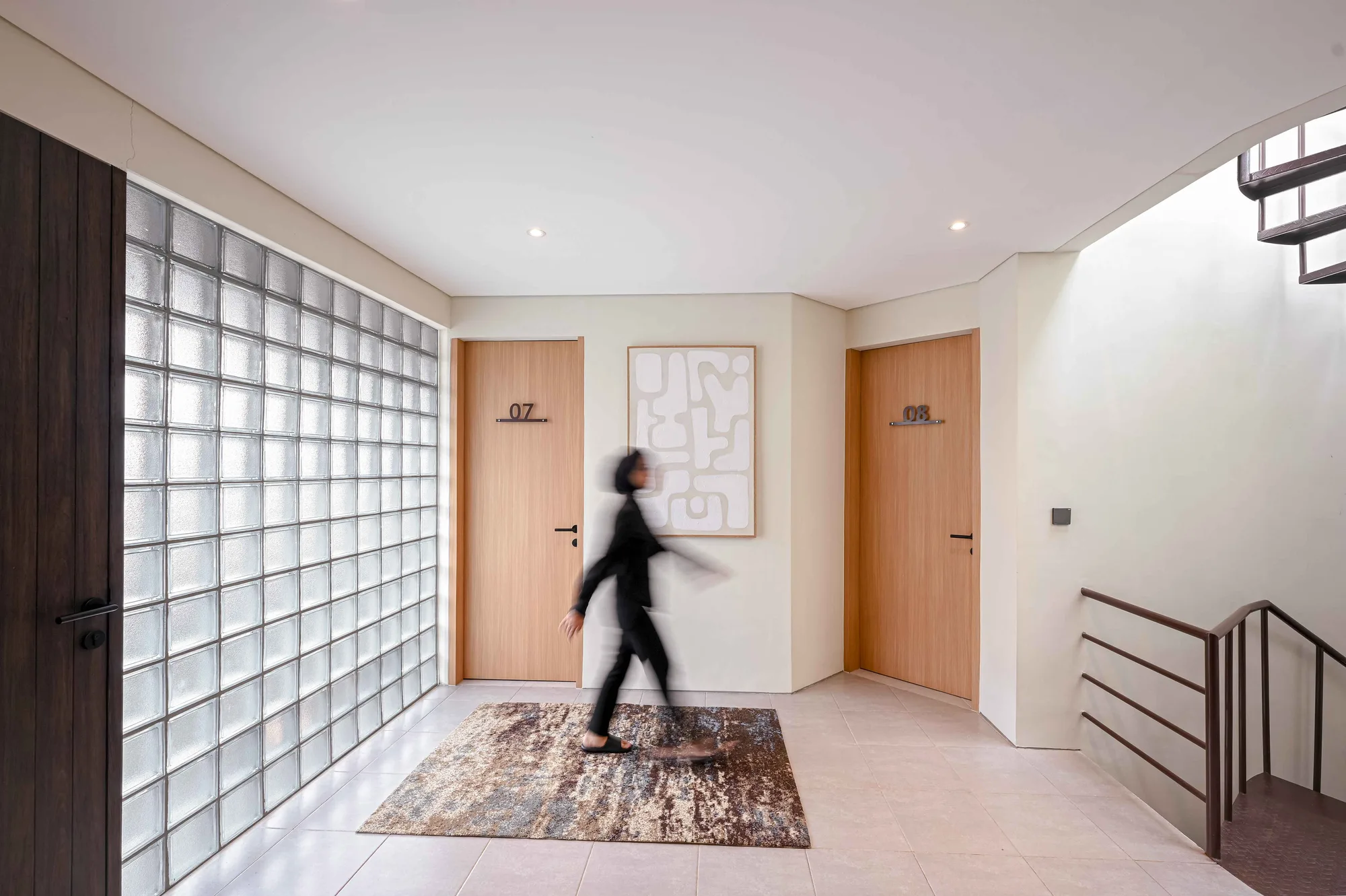 Hallway with rooms 07 and 08 either side of an abstract artwork, a resident crossing the rug in front of the full-height glass-block wall.