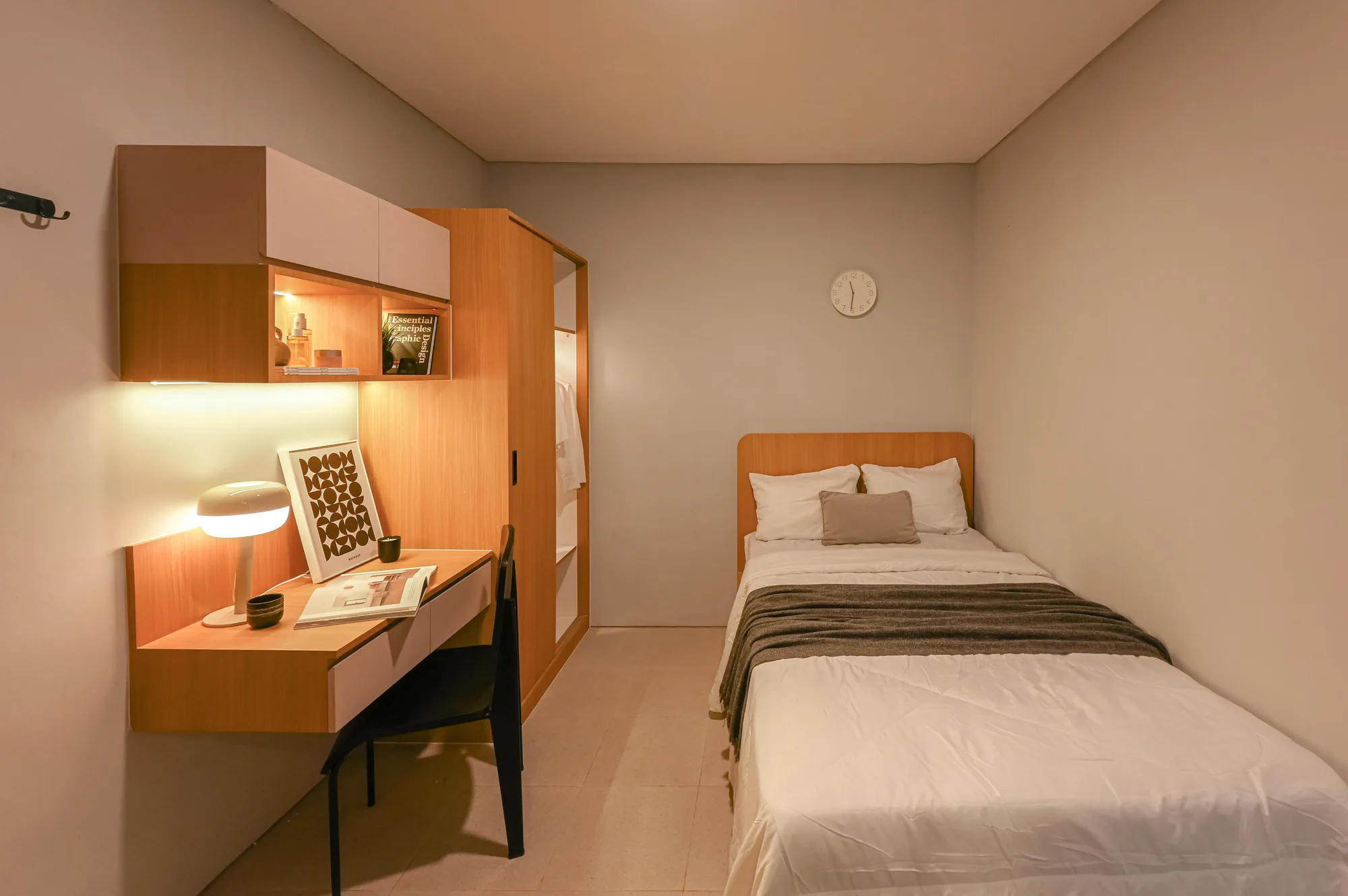 A guest room with a single bed against the wall, a wall clock above the headboard, and a built-in desk and wardrobe warm with wood veneer.