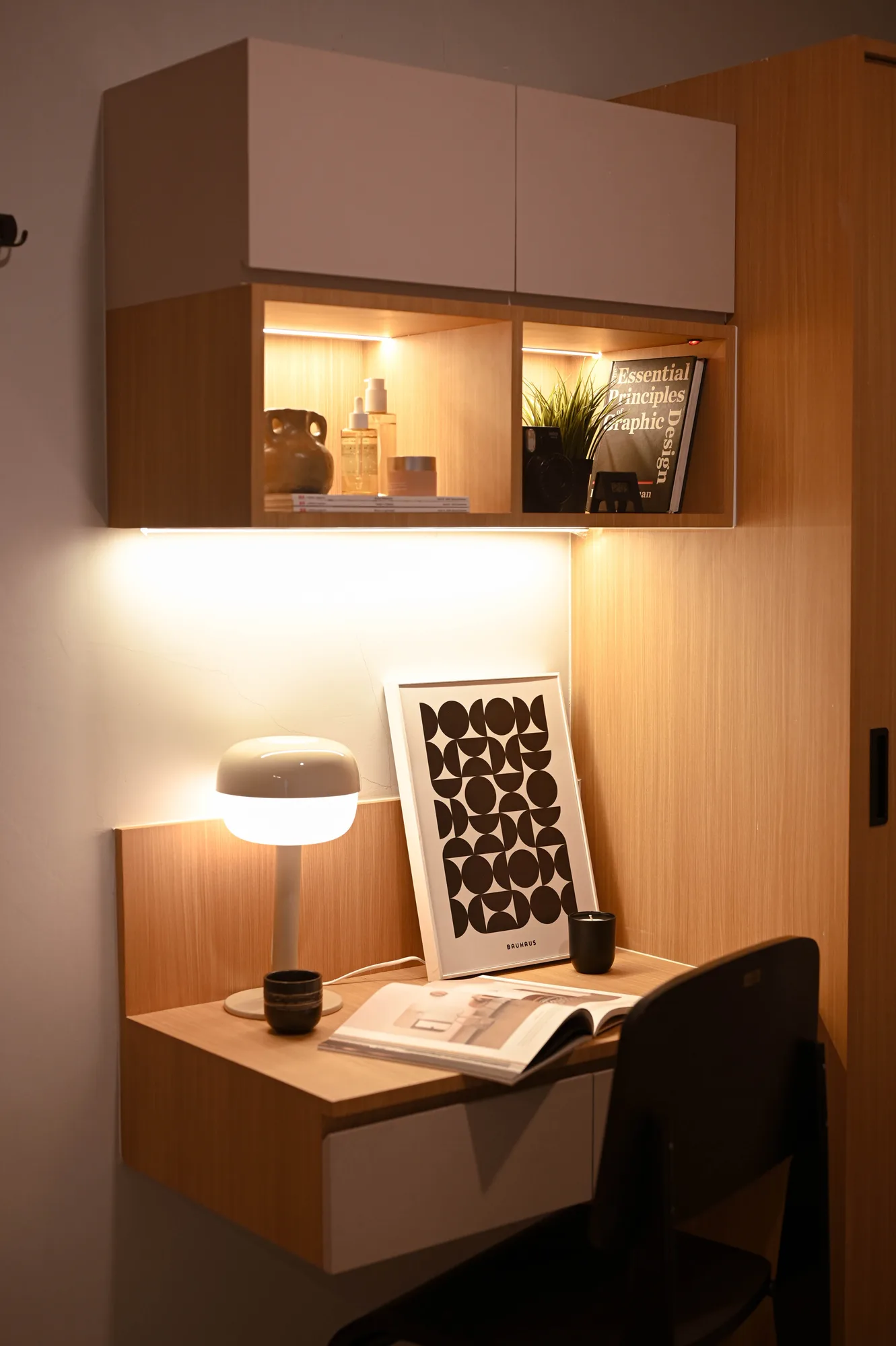 Built-in desk nook with a lit shelf of books and objects above, a lamp and framed print set against warm wood cabinetry.
