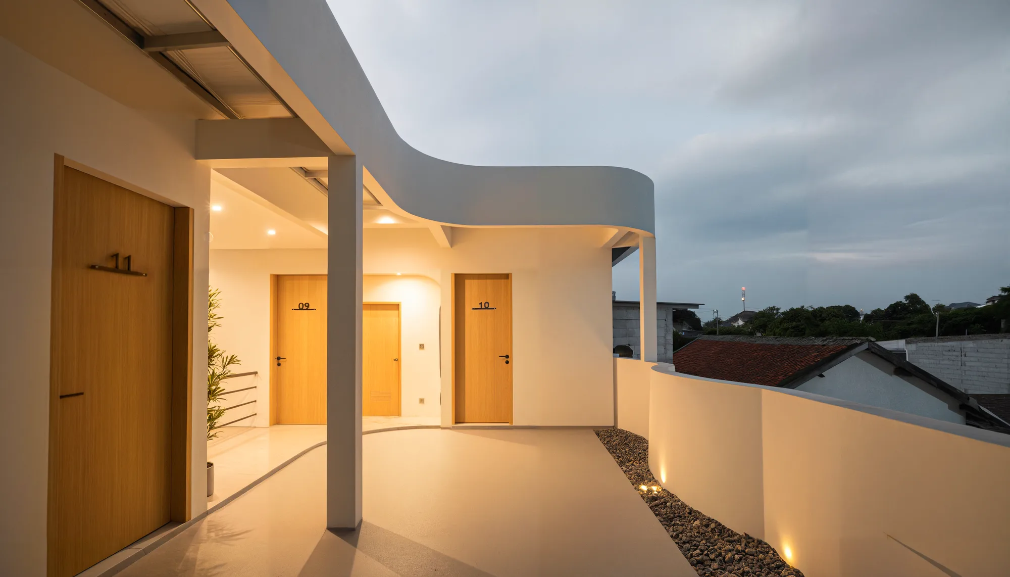 Dusk view along the rooftop corridor past rooms 09, 10 and 11, the curved parapet opening onto the surrounding neighborhood roofs.