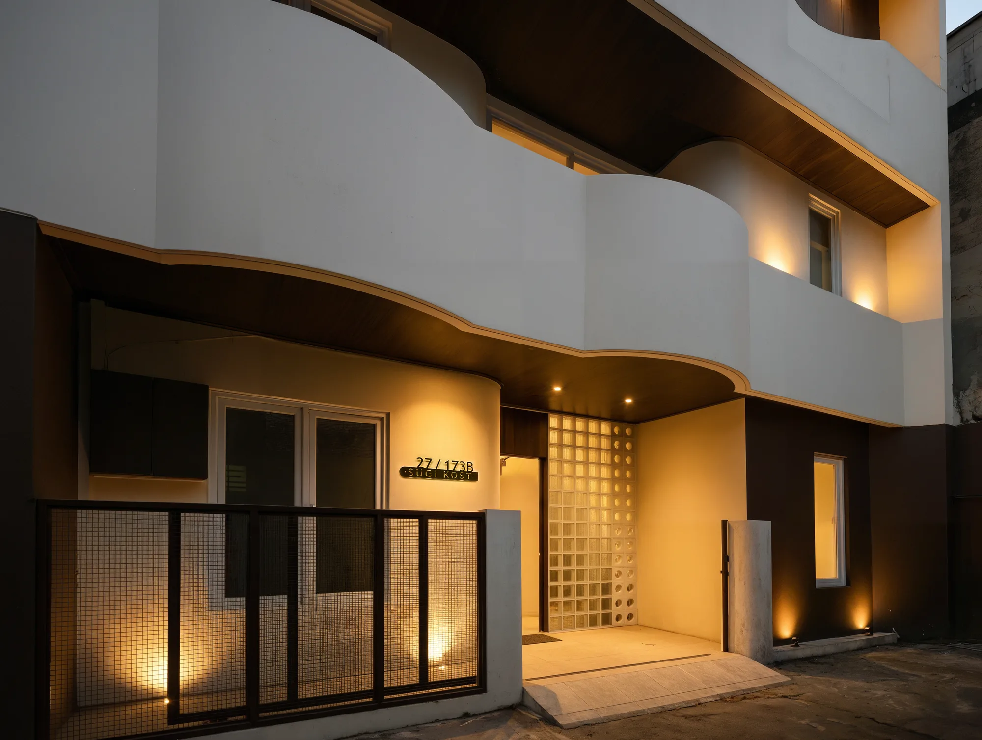 Sugi Kost's front facade lit at night, warm light spilling from the glass-block entrance through the gated courtyard.